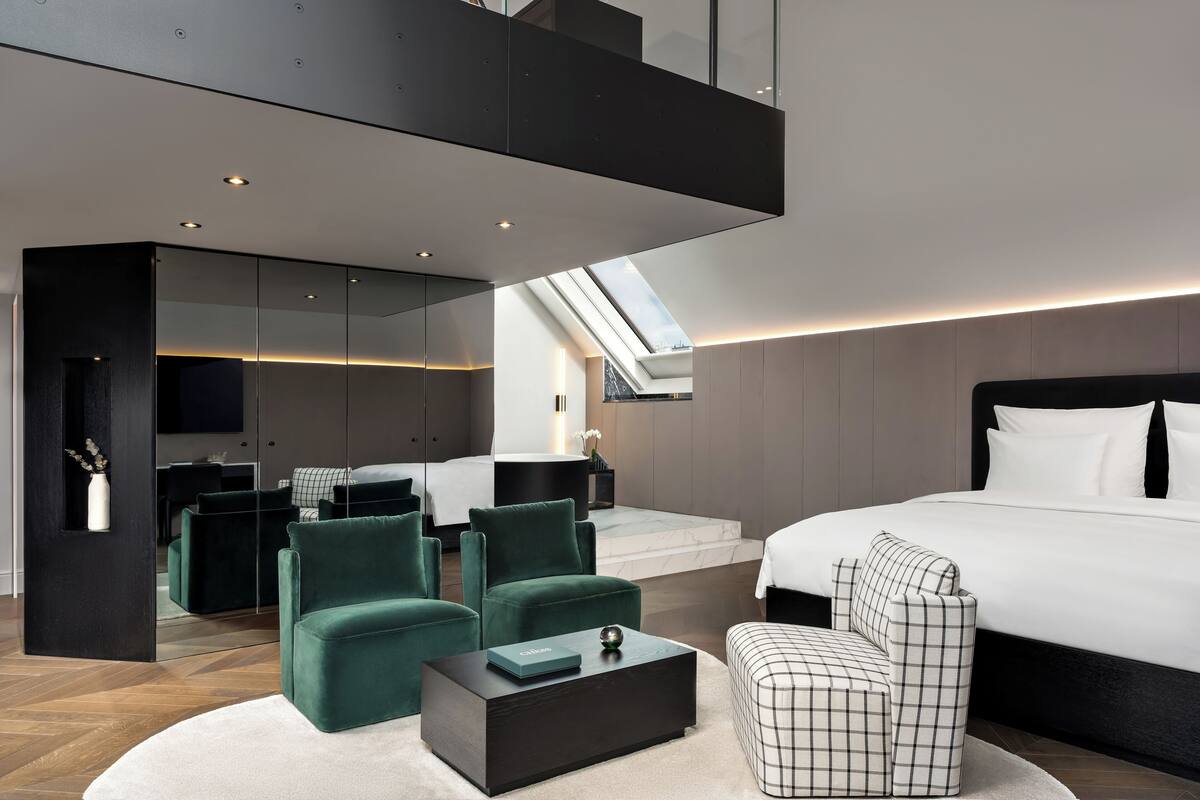 A modern bedroom with a large bed, two green armchairs, a checkered ottoman, a dark coffee table, and a skylight above the bed.