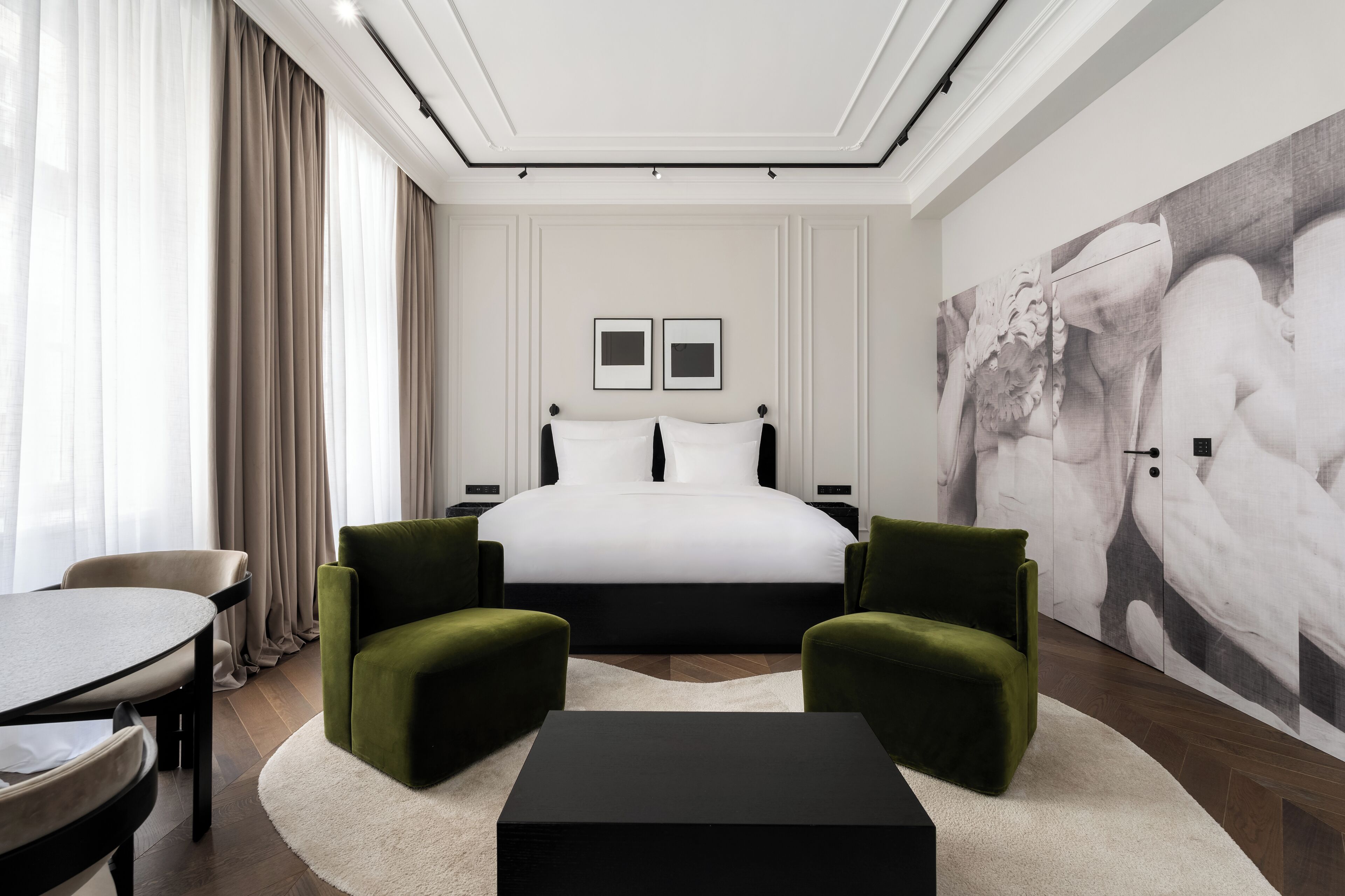 A modern hotel room with a large bed, two green armchairs, a black coffee table, a dining table, and a large abstract painting on the wall.