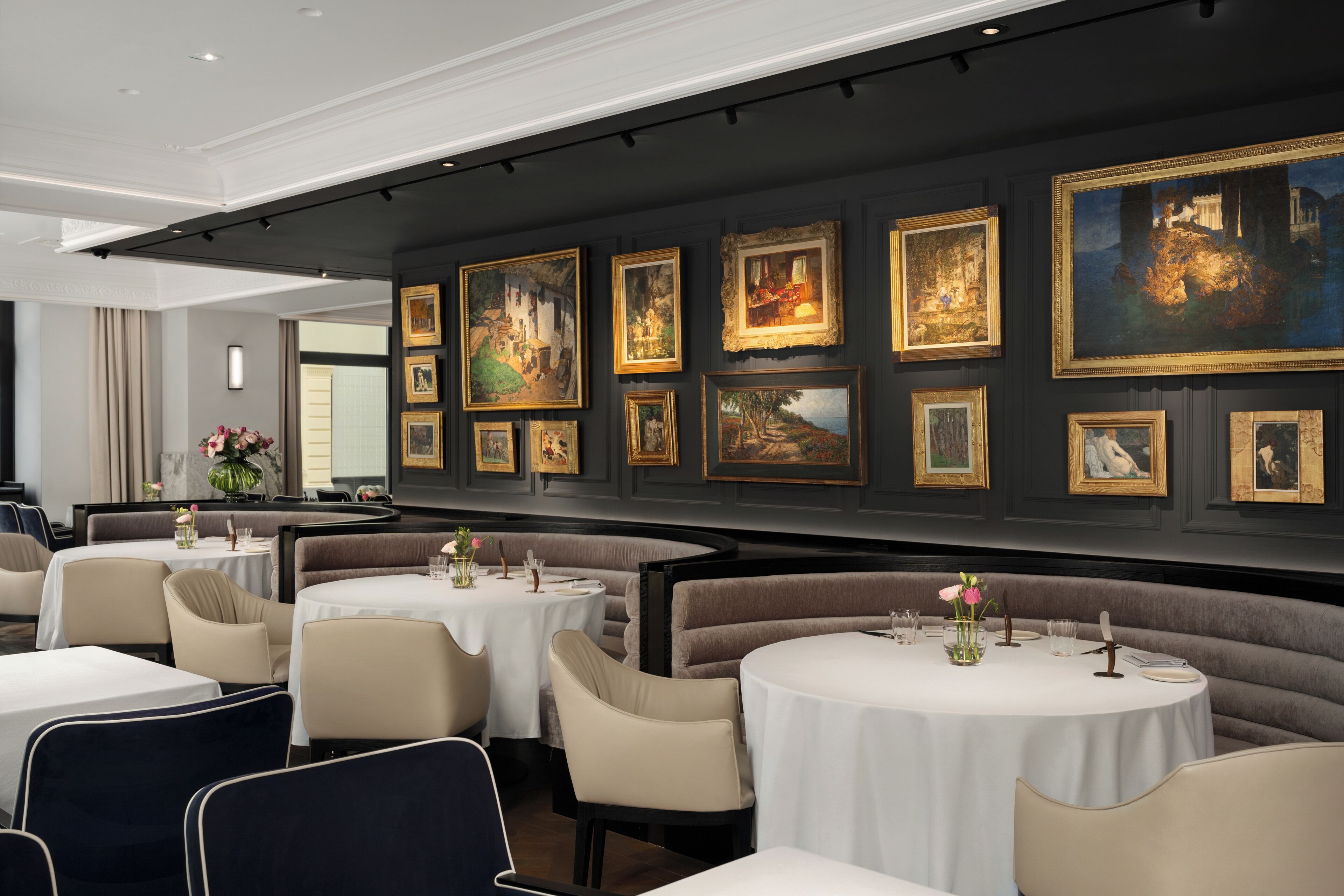 A restaurant with round tables, beige chairs, and a wall adorned with framed paintings.
