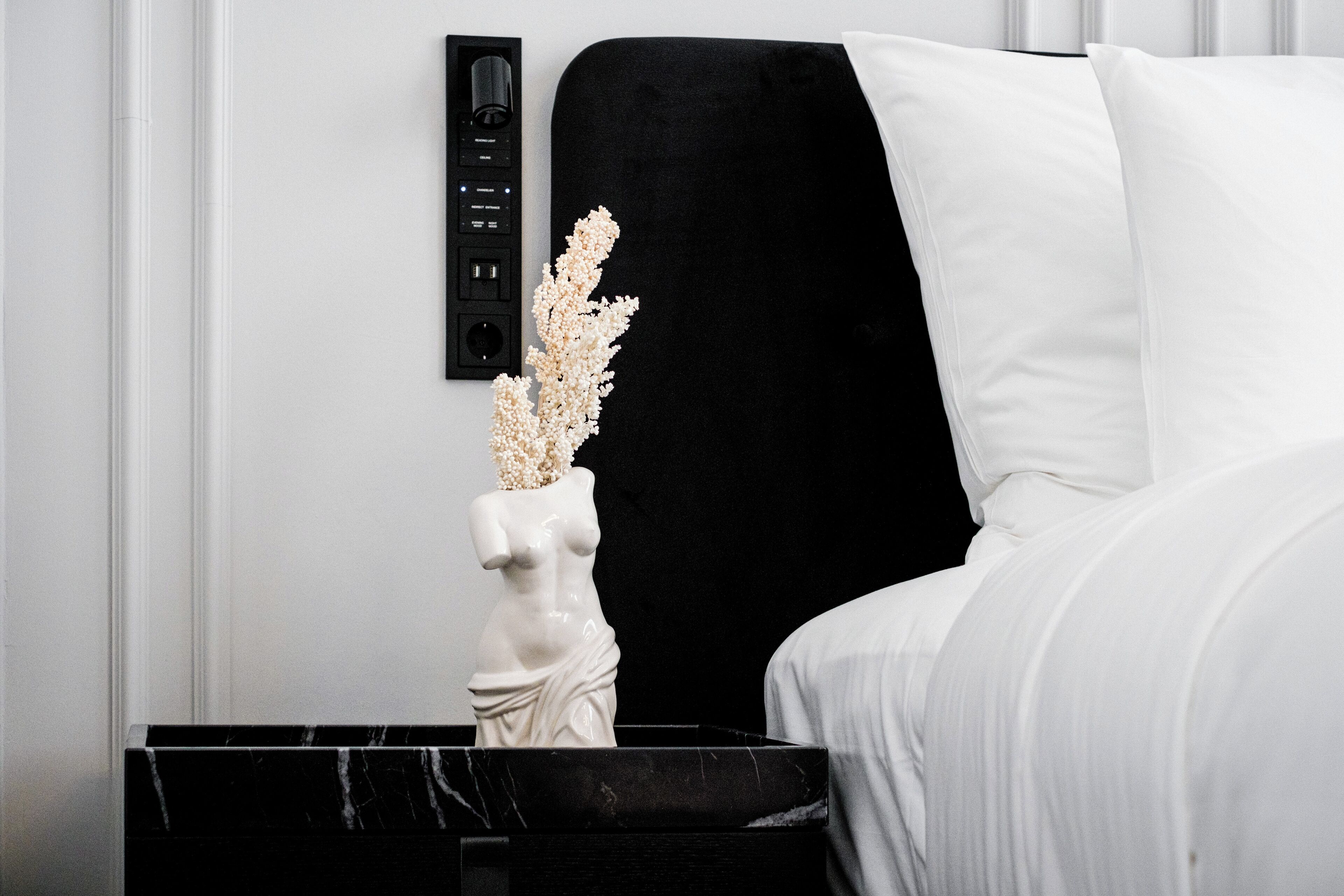A modern room with a black marble side table, a white vase with dried flowers, and a white upholstered headboard.