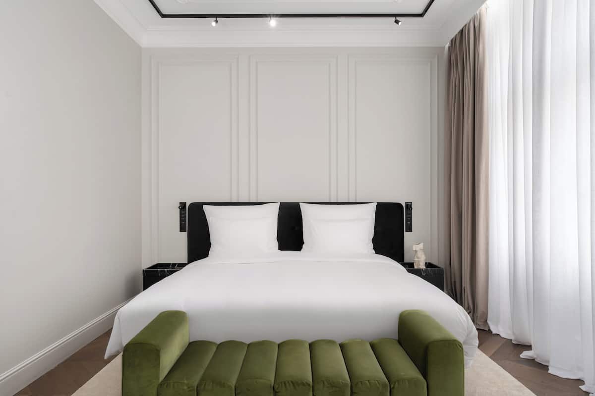 A modern hotel room with a large bed, a green bench, and a nightstand with a lamp.