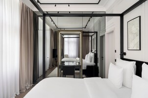A modern hotel room with a large bed, a desk, and a chair. There is a mirror on the wall and a window with curtains.