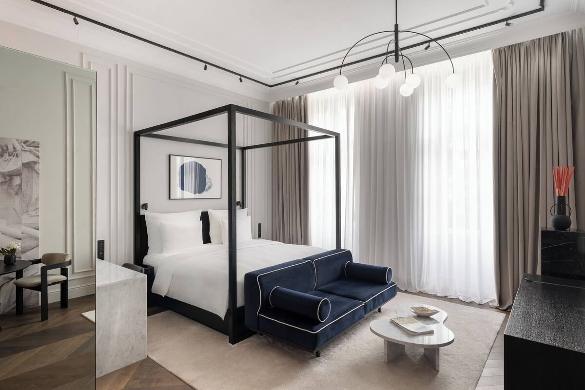 A modern hotel room with a four-poster bed, a blue sofa, a small round table, and a marble coffee table.