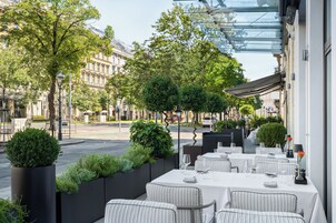 An outdoor dining area with tables set for two, surrounded by greenery and overlooking a street with buildings.