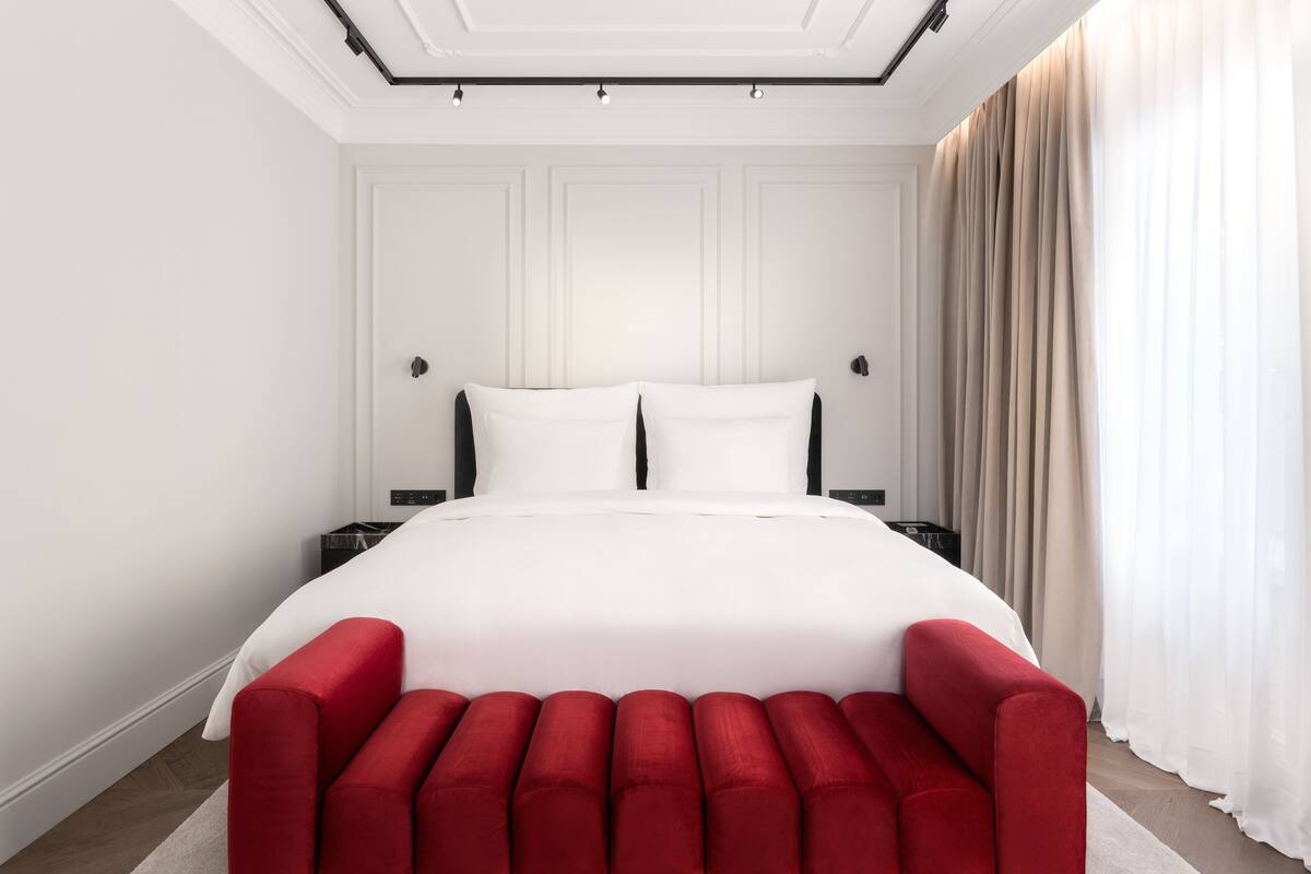 A large bed with white linens, a red upholstered bench, and a nightstand with a lamp.