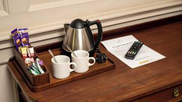 A wooden tray with a kettle, two white mugs, a remote control, and a box of chocolates on a wooden surface.