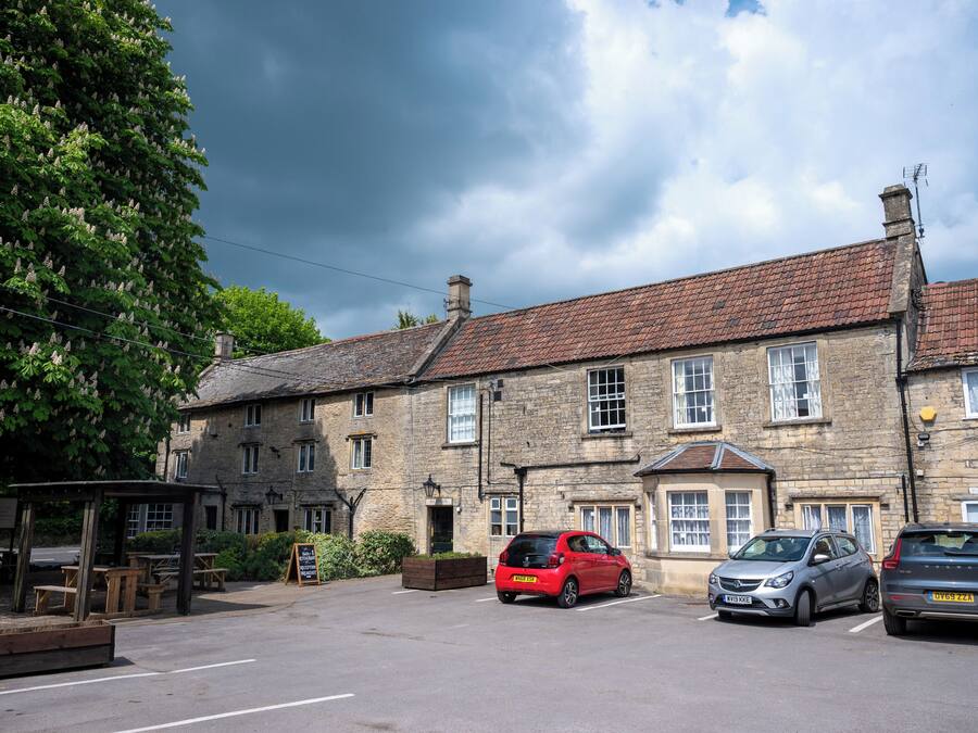 A stone building with a tiled roof, a parking area with cars, and a wooden shelter with benches.