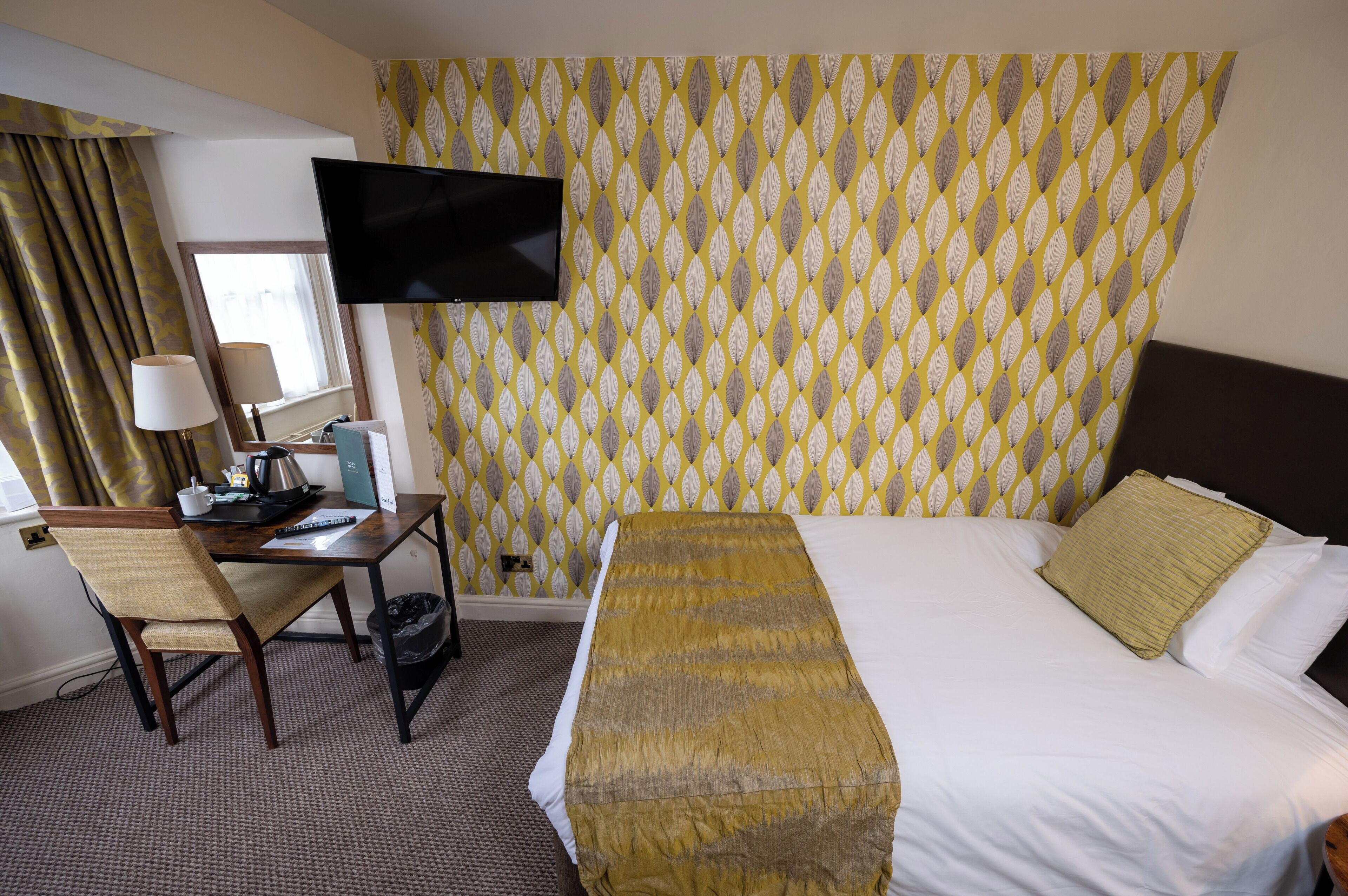 A hotel room with a bed, a desk with a chair, a television, and a patterned wallpaper.