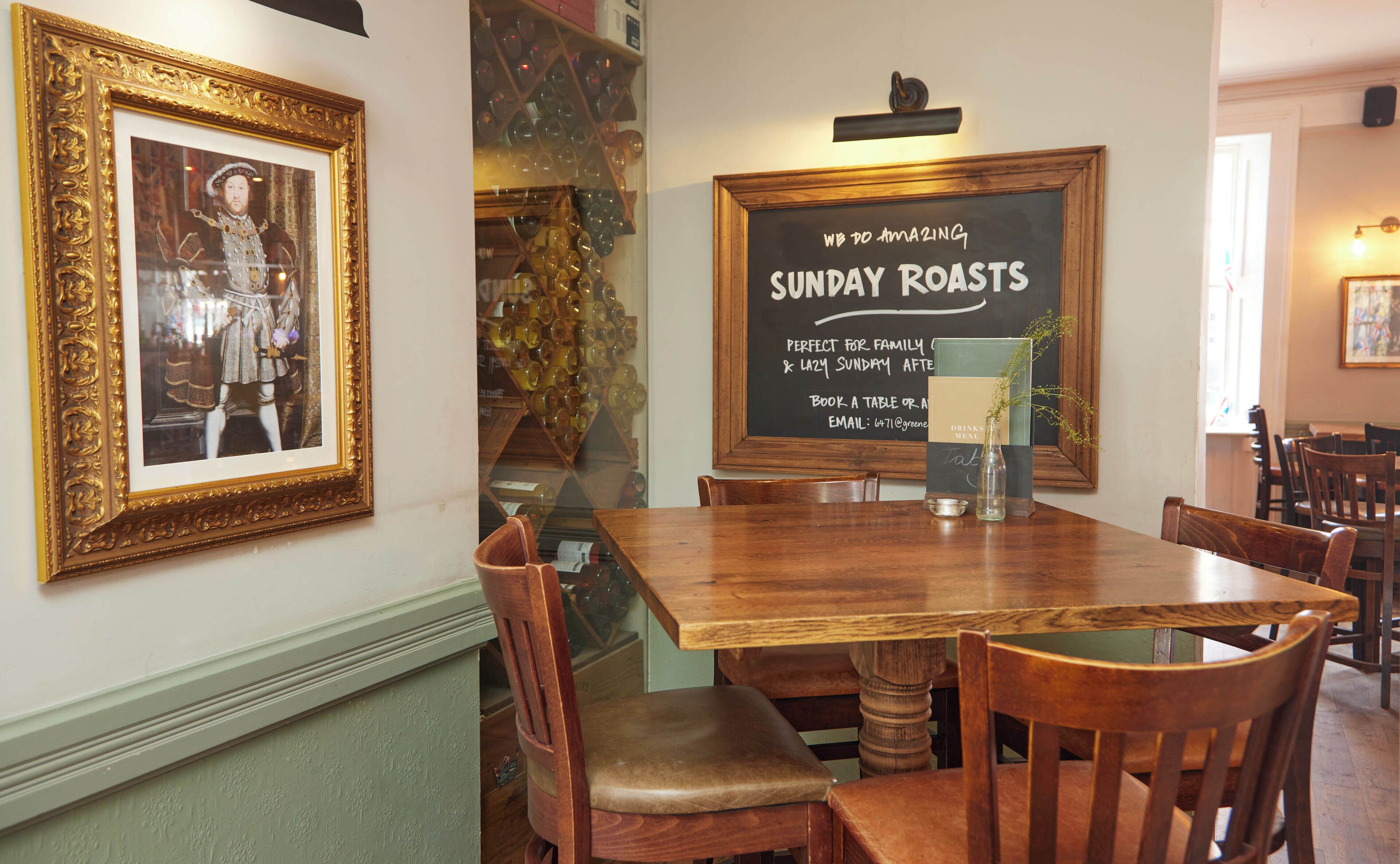 A dining area with a wooden table and chairs, a framed picture on the wall, and a chalkboard sign advertising Sunday roasts.