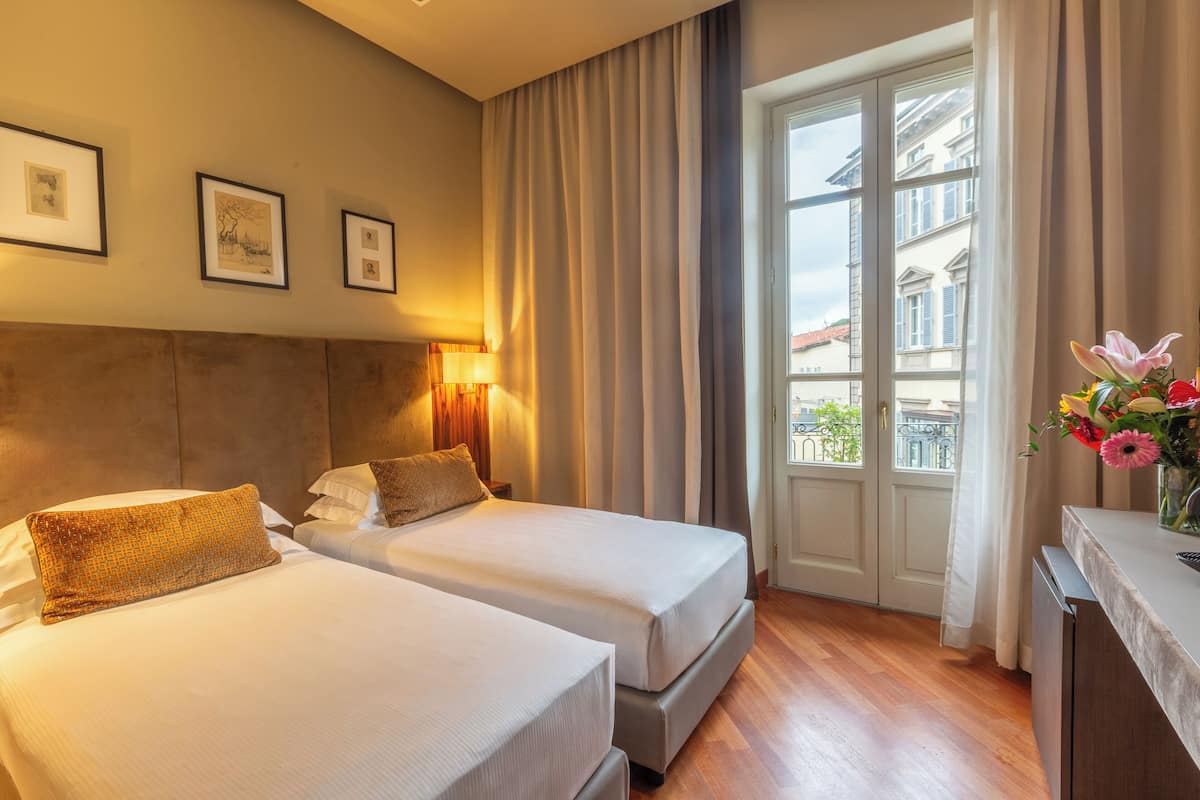 A hotel room with two beds, a window with curtains, a desk with a vase of flowers, and framed pictures on the wall.
