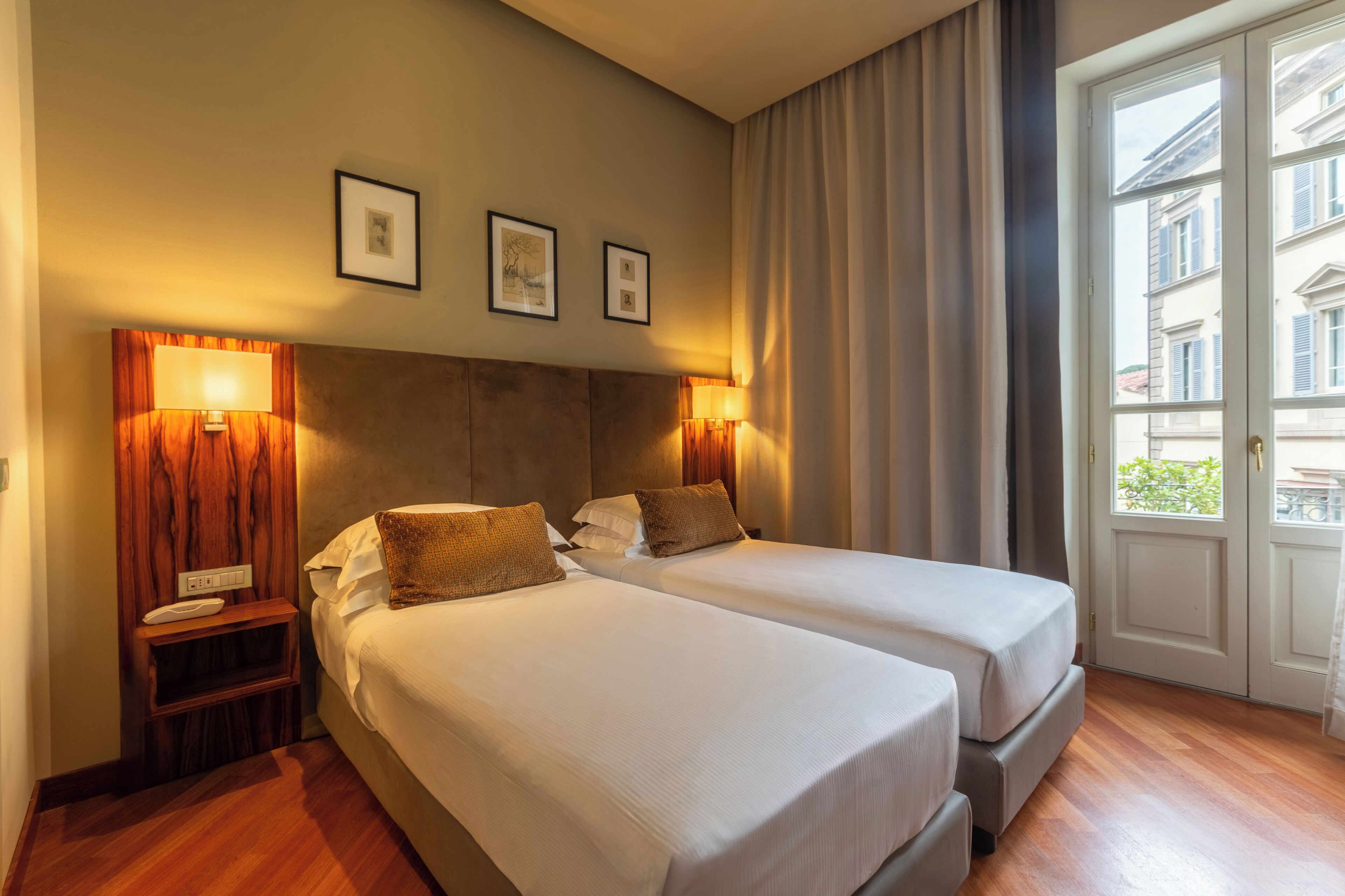 A hotel room with two beds, a wooden headboard, bedside lamps, a small table, and a window with curtains.
