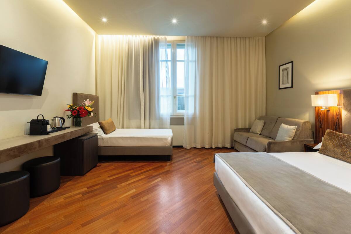 A hotel room with a bed, a sofa, a desk with a flat-screen TV, a coffee maker, and a window with curtains.