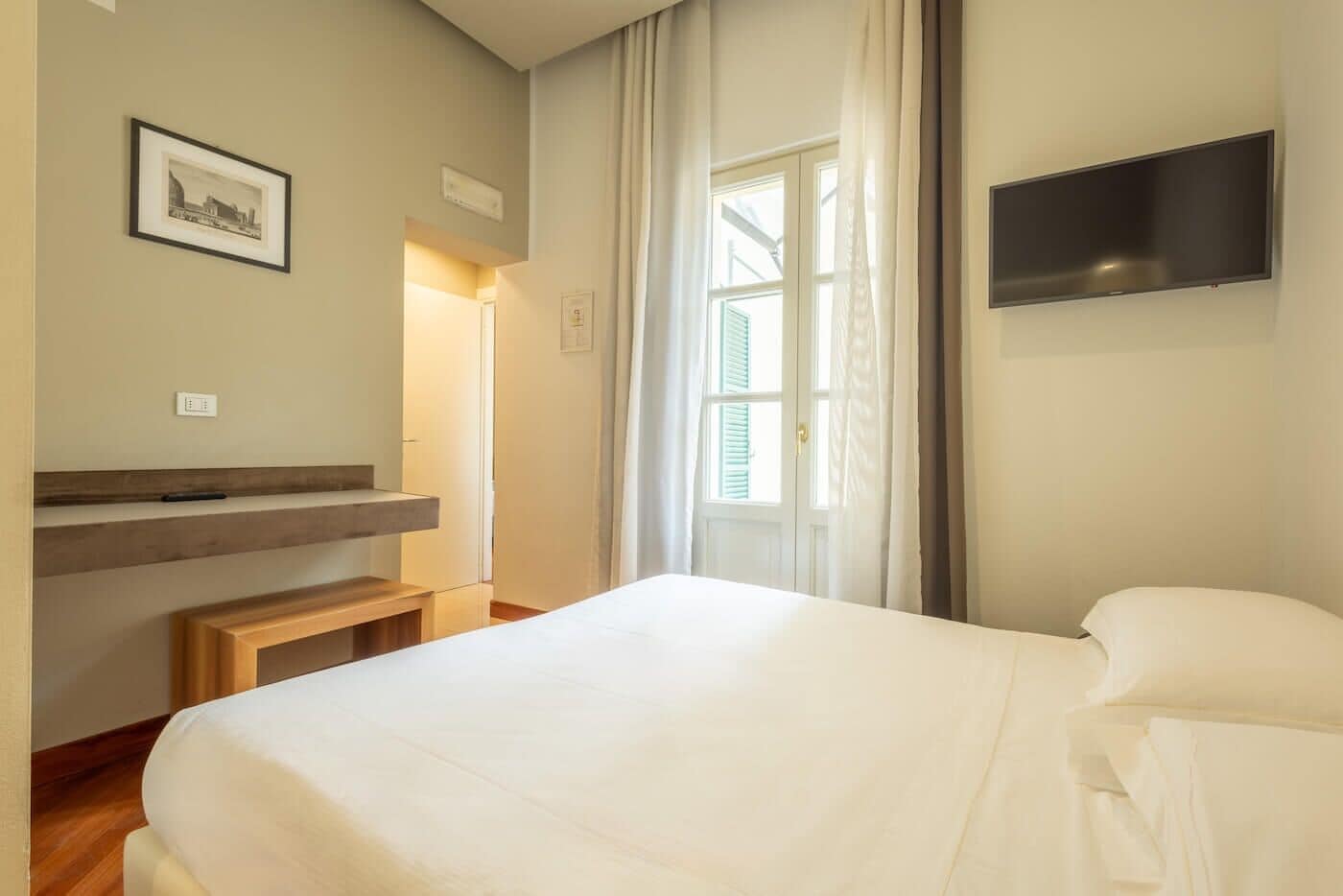 Una camera d'albergo con un letto, una TV a parete, una piccola panca in legno, un quadro appeso e una finestra con tende.