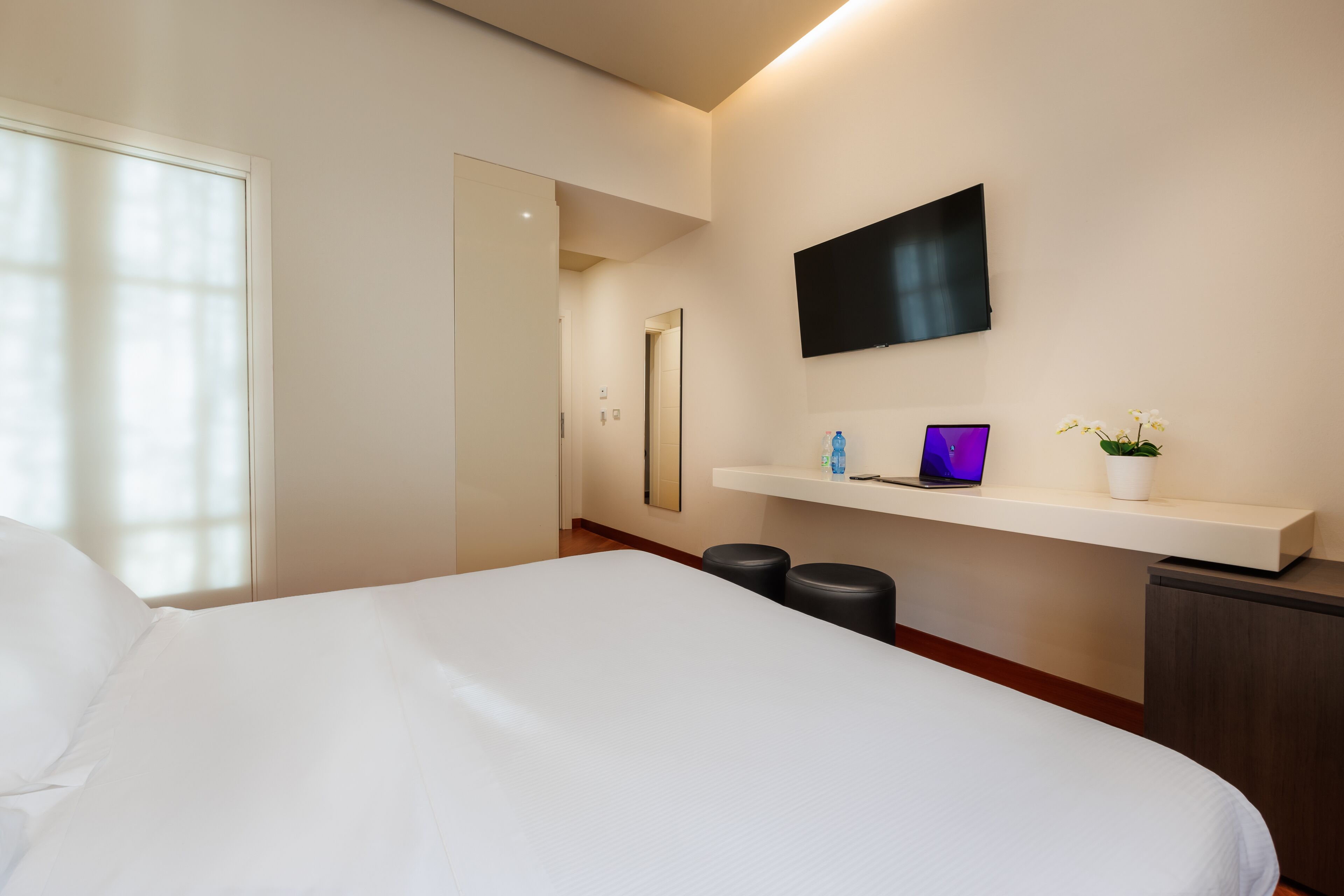Una camera d'hotel moderna con un letto grande, una TV a parete, una piccola scrivania con un portatile e una pianta in vaso.