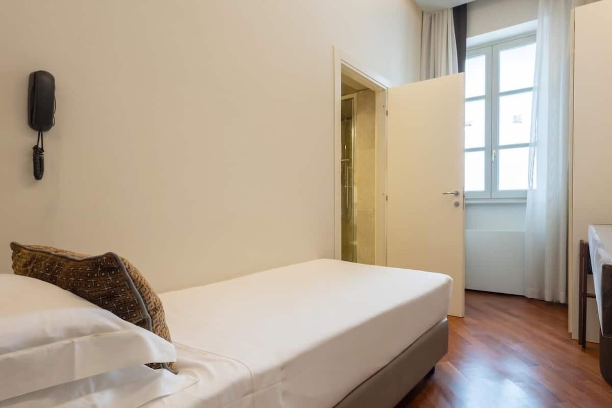 A single-bed room with a white mattress, a patterned pillow, a telephone on the wall, and a door leading to a bathroom.