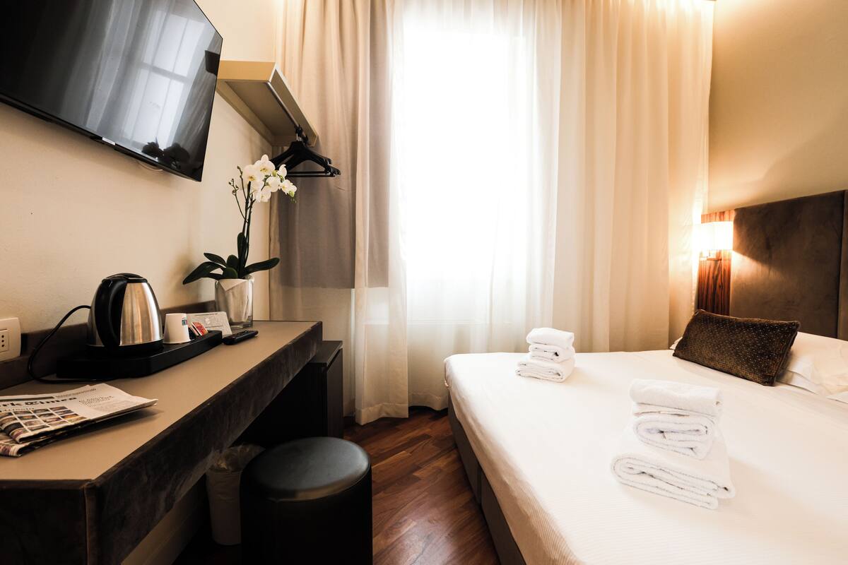A hotel room with a bed, a desk, a TV, a window with curtains, and a bedside lamp.