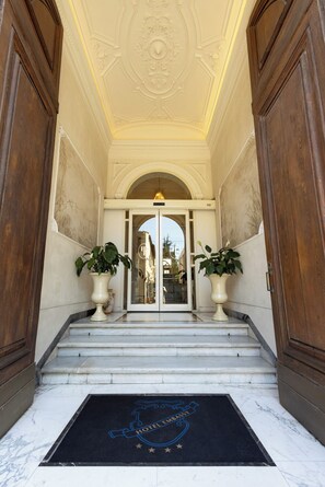 A grand entrance with marble steps, a black doormat with a hotel emblem, and two large potted plants. - Hotel Embassy (Florence)