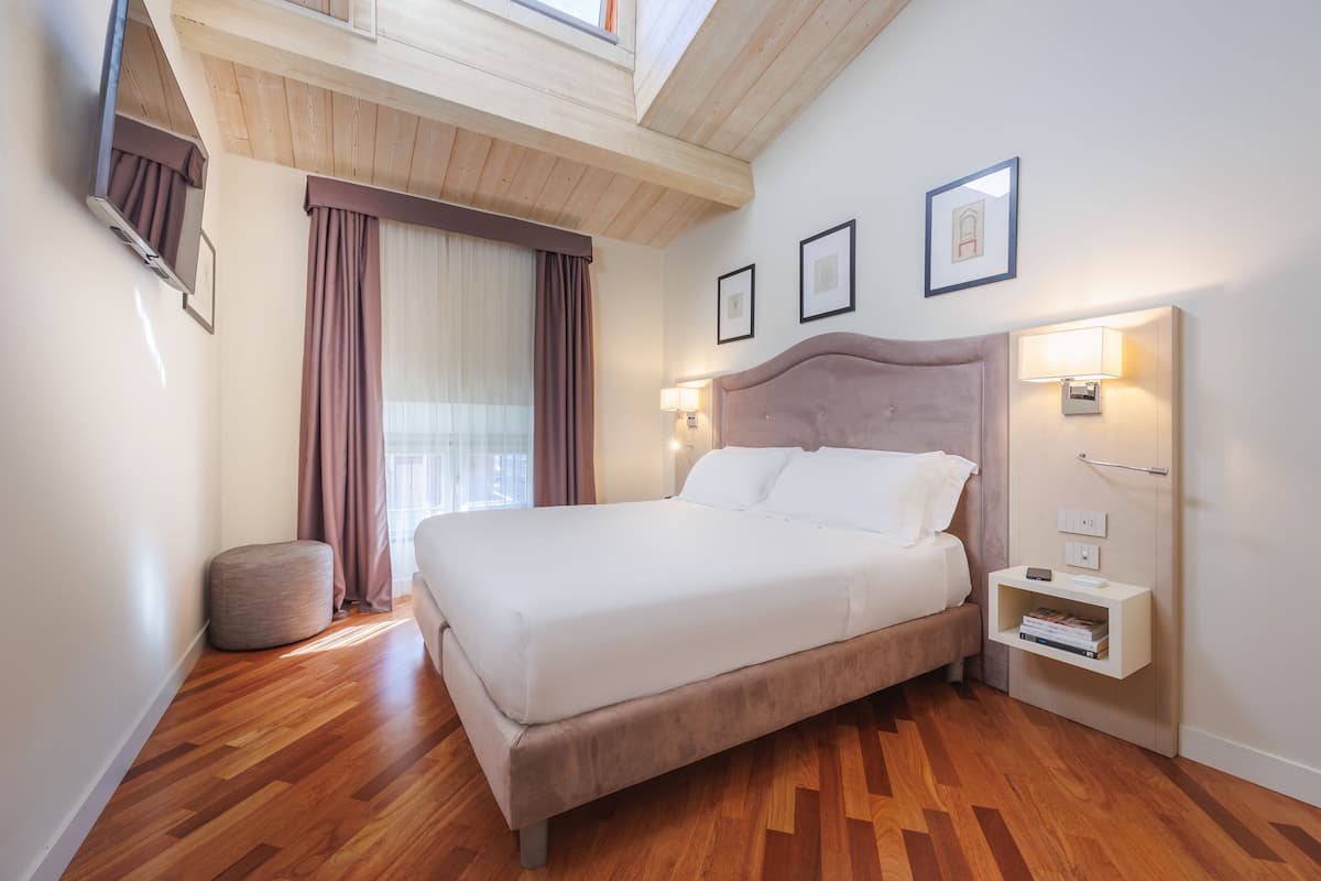 A neatly arranged bedroom with a large bed, wooden flooring, a bedside table with a lamp, and a window with curtains.