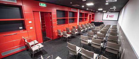 A conference room with rows of chairs, a projector screen, and a red exit sign.