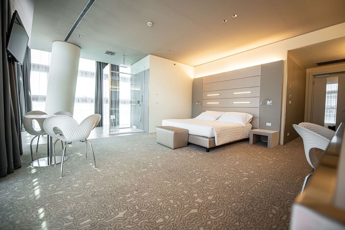 Moderni hotellihuone, jossa on suuri sänky, oleskelualue tuoleineen, lasiseinäinen suihkutila ja litteänäyttöinen televisio.
