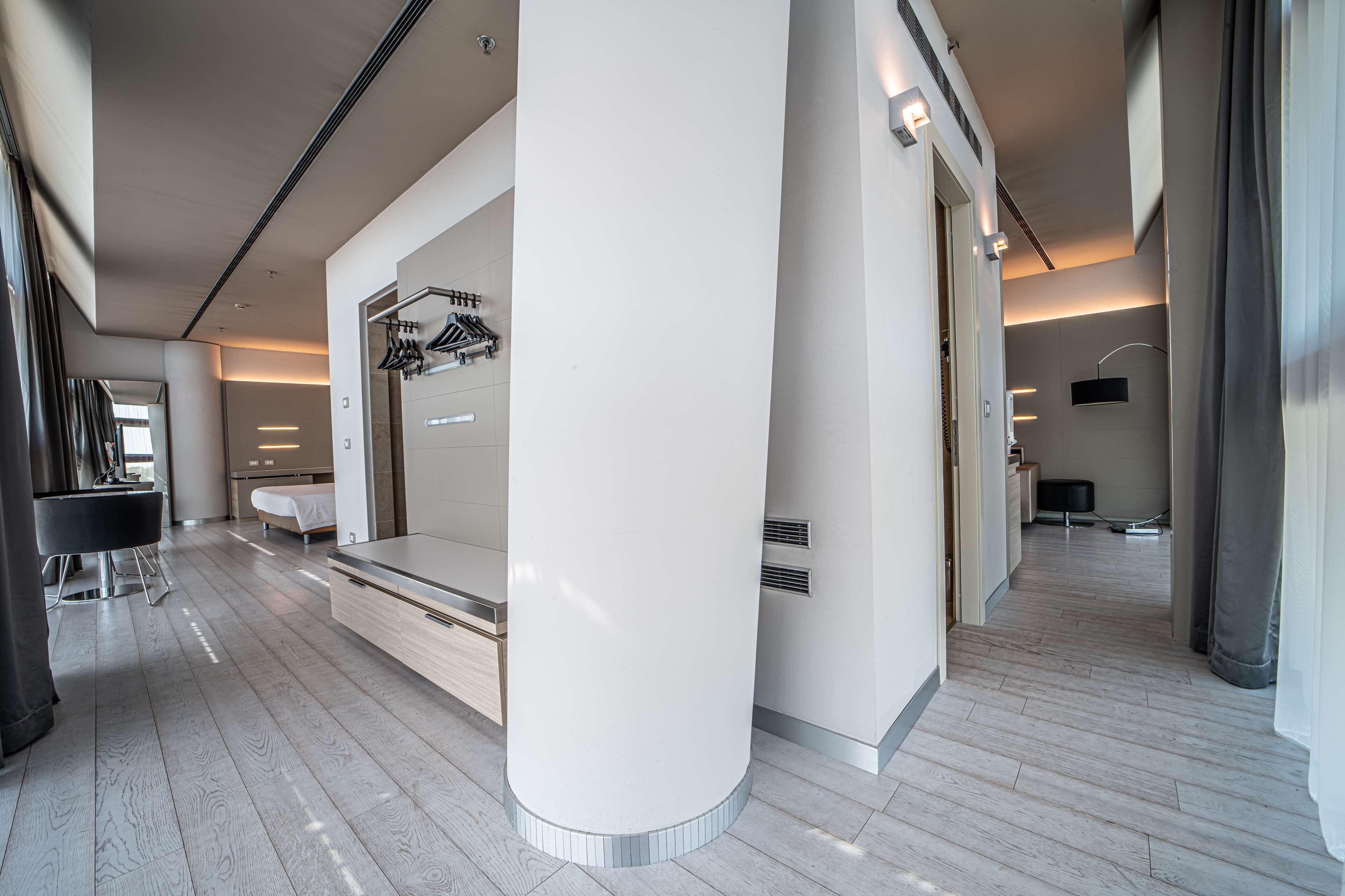 Habitación de hotel moderna con una cama grande, tocador con espejo, área de descanso con banco y pasillo que da a otras habitaciones.
