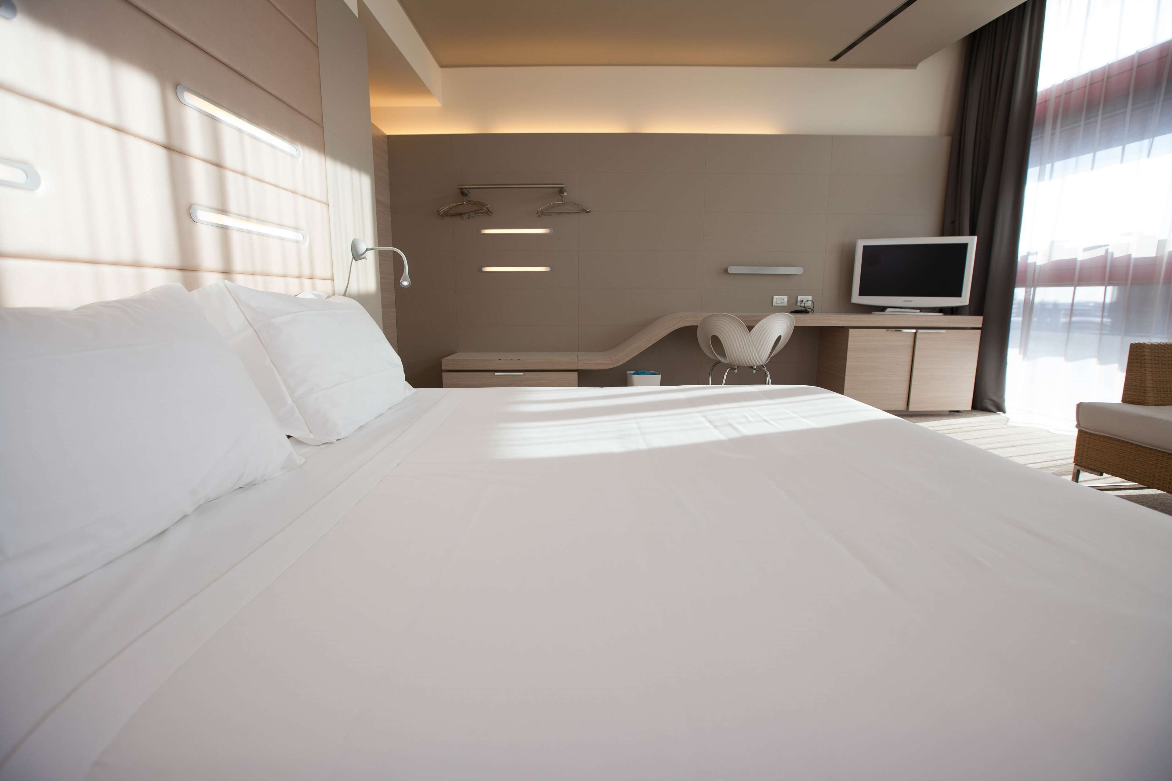 Una habitación de hotel moderna con una cama grande, televisor, zona de estar y ventana con cortinas.