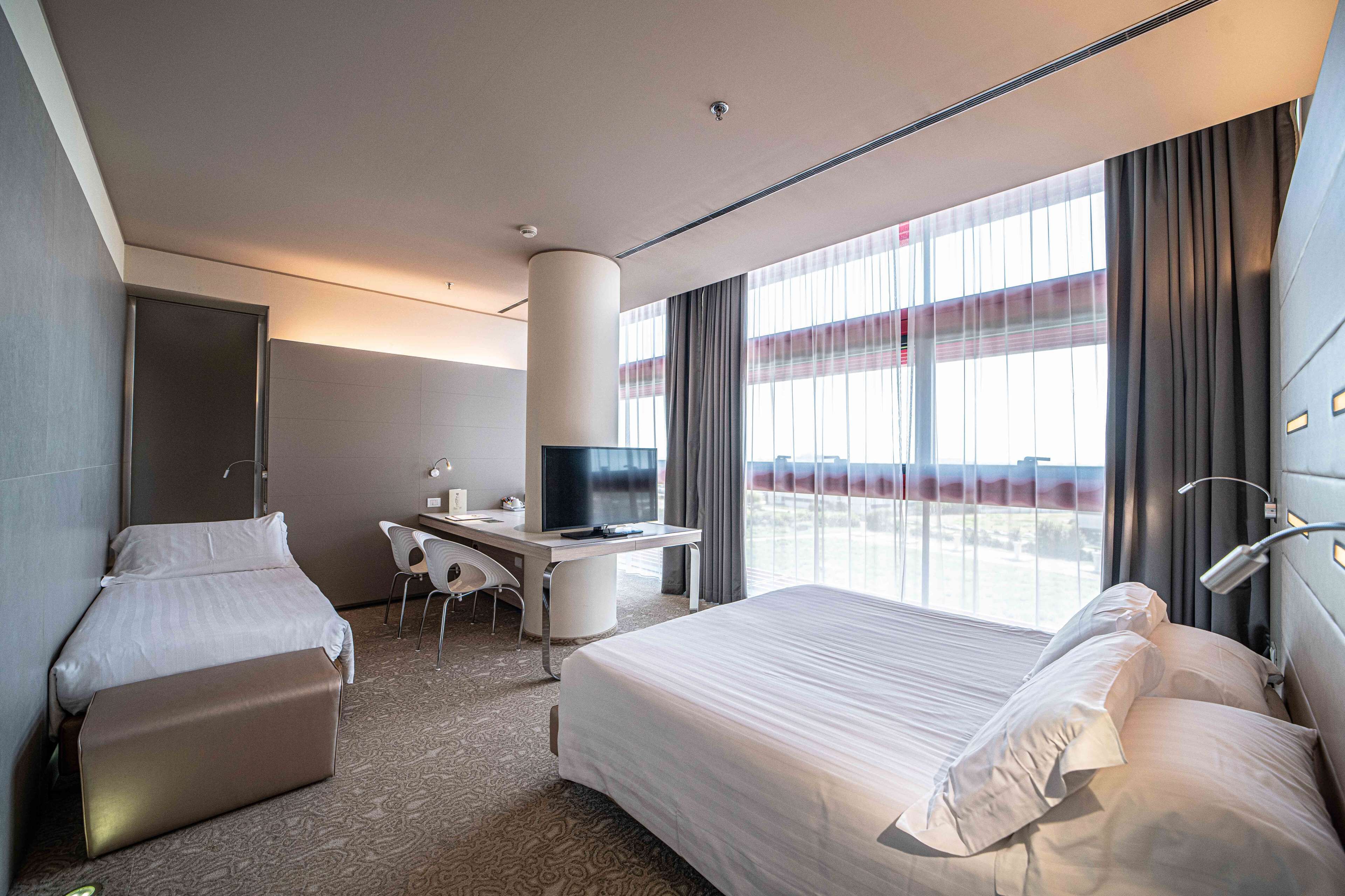 A hotel room with a bed, a desk, a chair, a TV, and large windows with curtains.