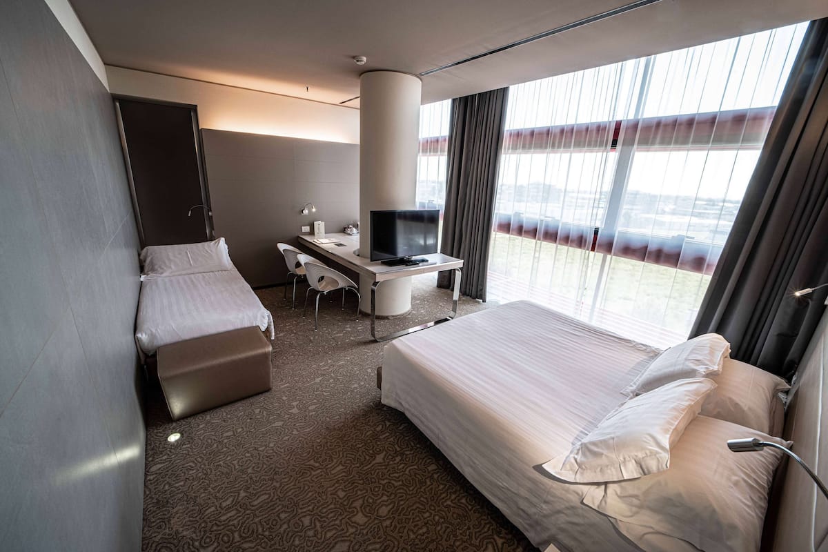 Hotellihuone, jossa on sänky, työpöytä, tuoli, televisio ja suuri ikkuna, jossa on verhot.