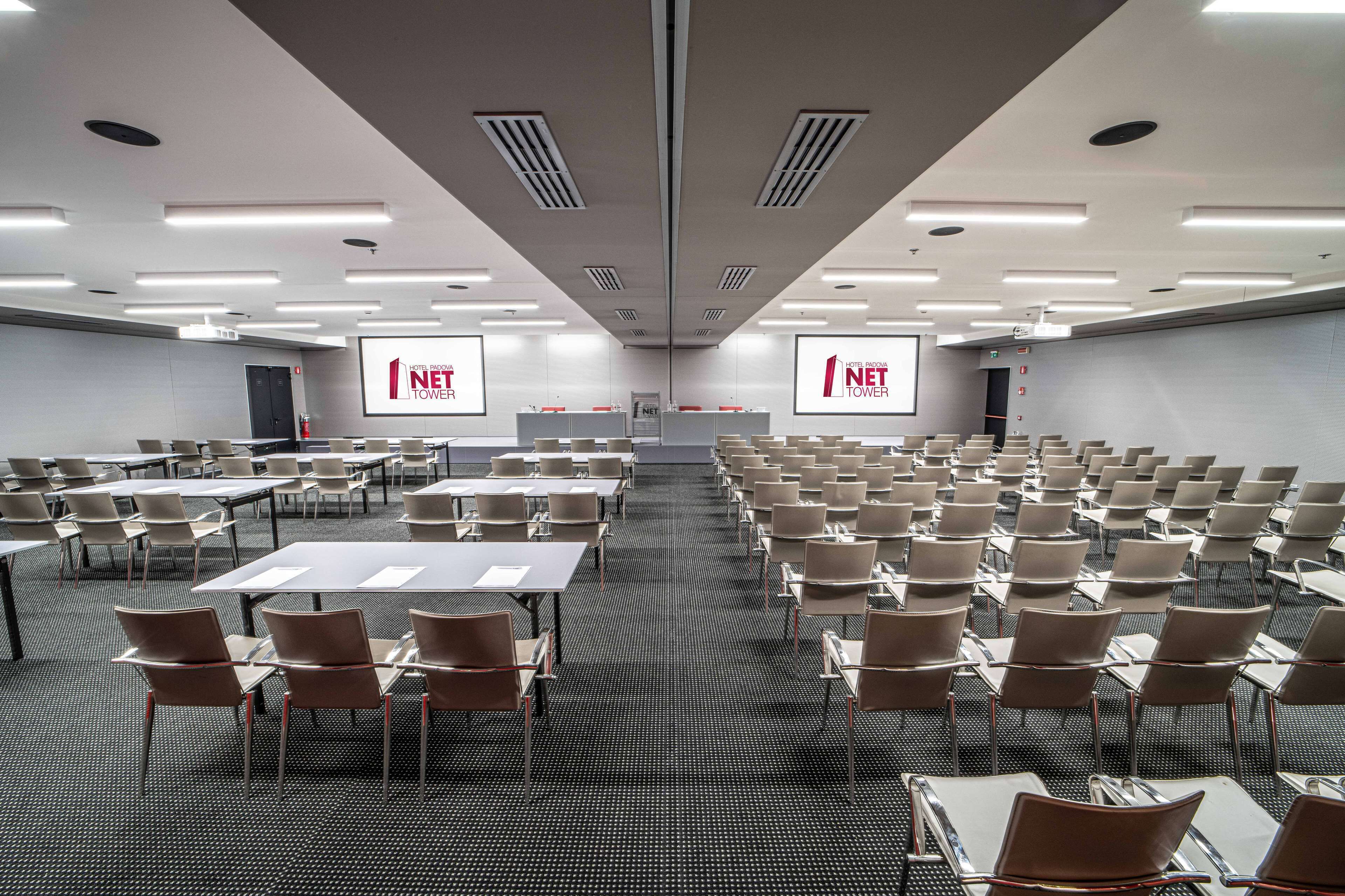 Un salón de conferencias con filas de mesas y sillas, una pantalla grande mostrando '1 NET TOWER' y un diseño de techo moderno.