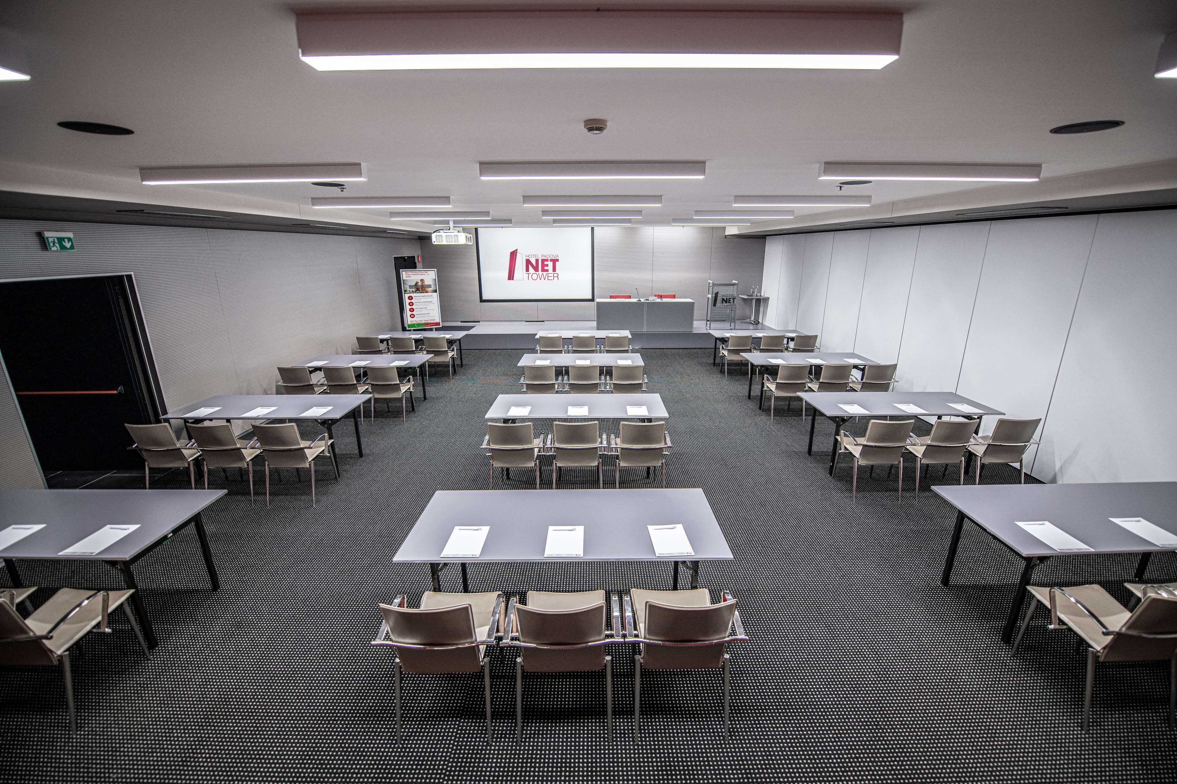 A conference room with multiple tables and chairs, a projector screen displaying 'NET TOWER', and a presentation podium.