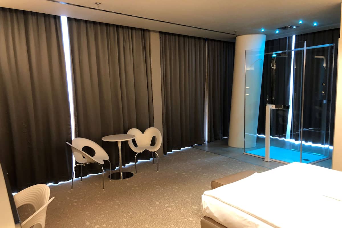 Hotellihuone, jossa on suuri sänky, pieni pyöreä pöytä, kaksi valkoista tuolia ja suuri ikkuna, jossa on verhot.