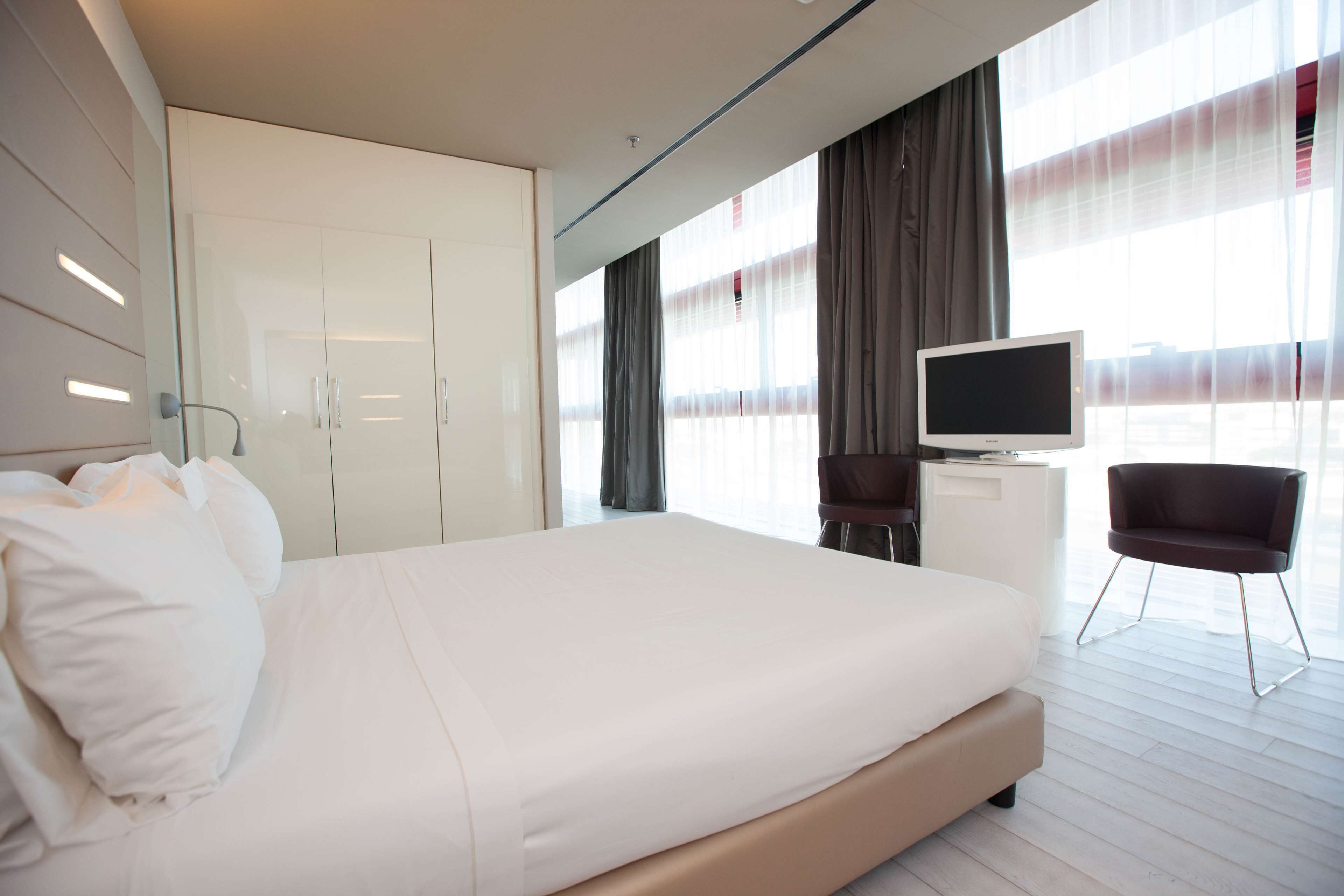 Camera d'albergo moderna con un letto grande, una TV su un mobile bianco, una sedia e ampie finestre con tende.