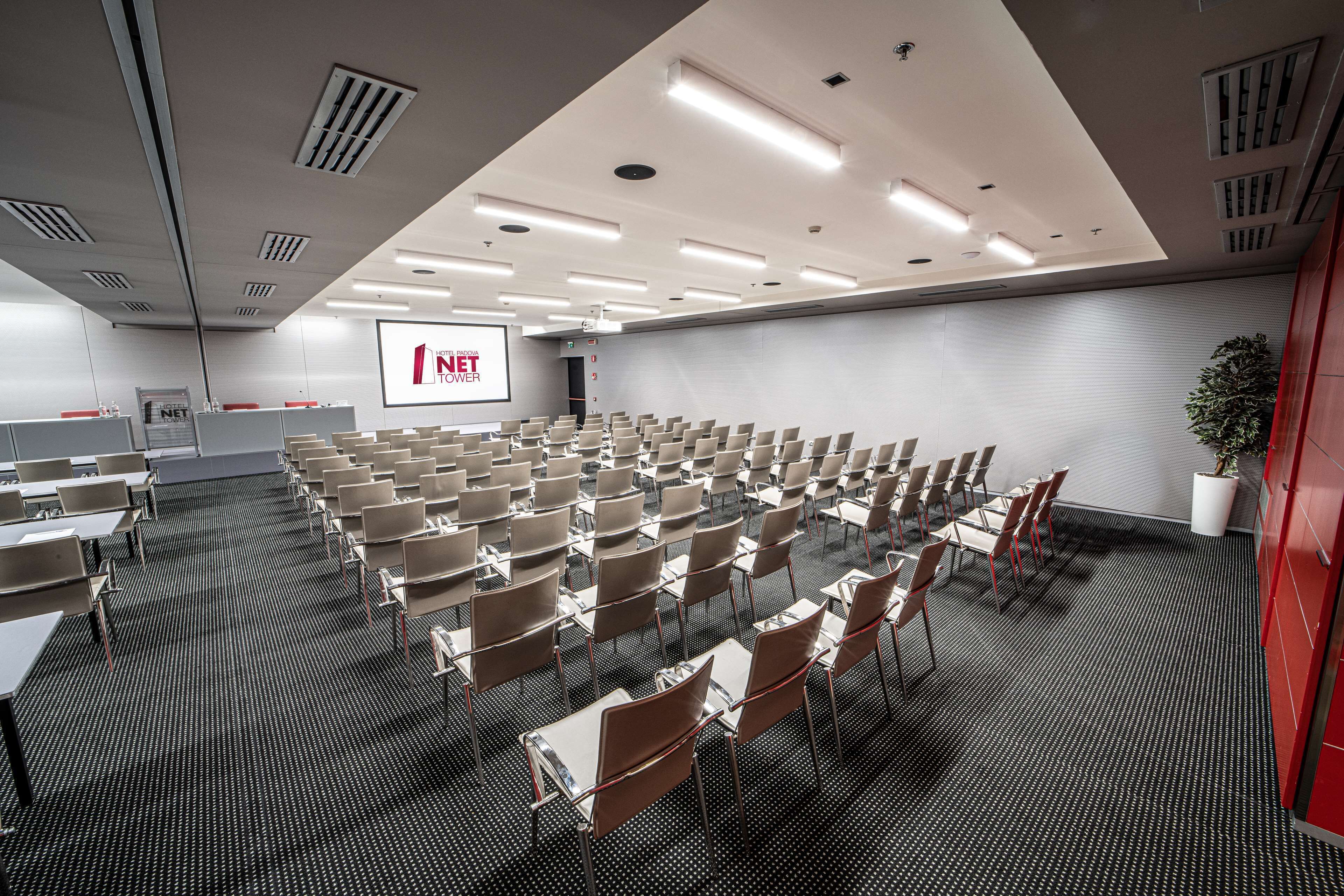 Un salón de conferencias con filas de sillas y mesas, una pantalla de proyector y una pizarra.
