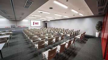 Un salón de conferencias con filas de sillas y mesas, una pantalla de proyector y una pizarra.