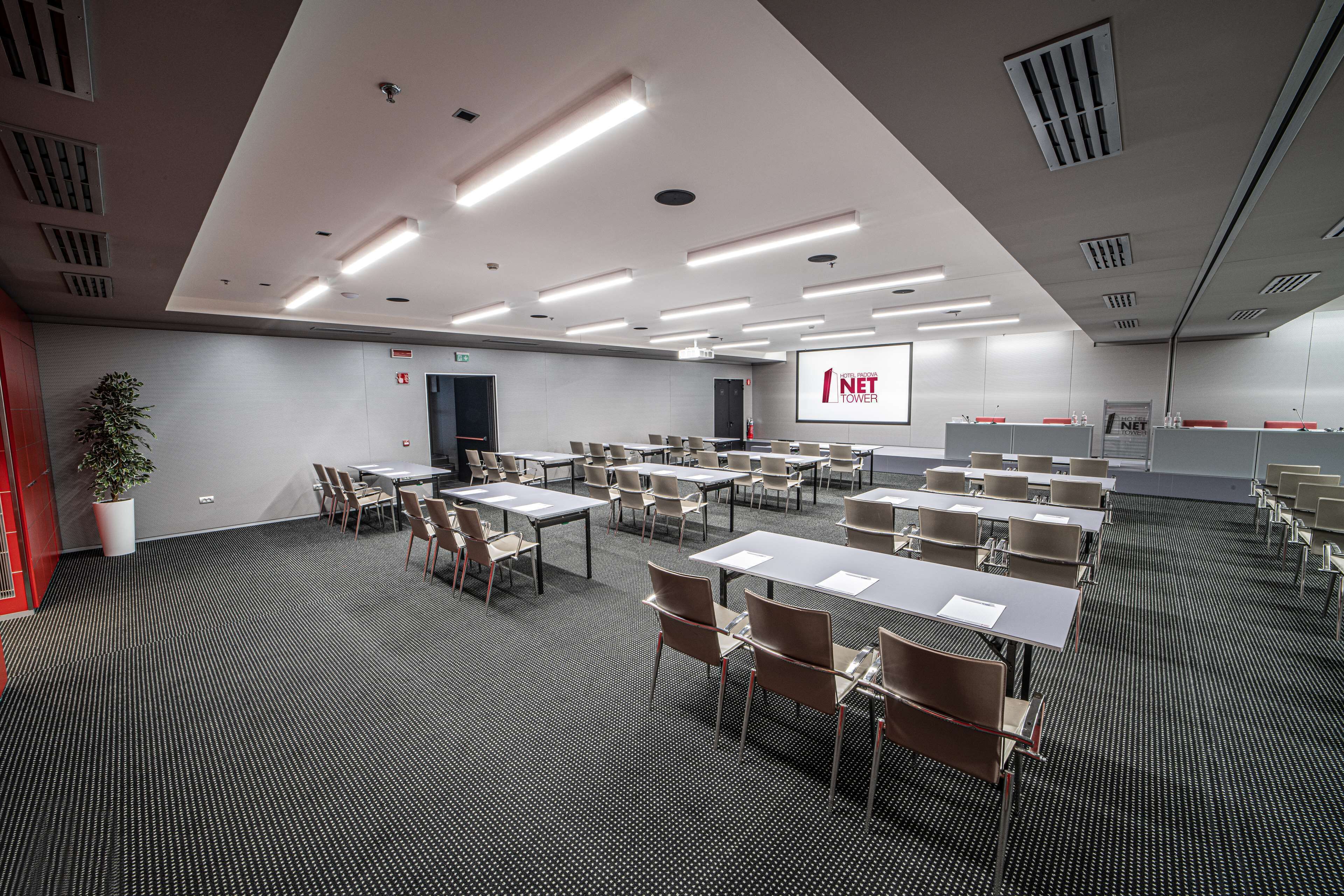 Une salle de conférence équipée de tables et de chaises, d’un écran de projection et d’une plante.