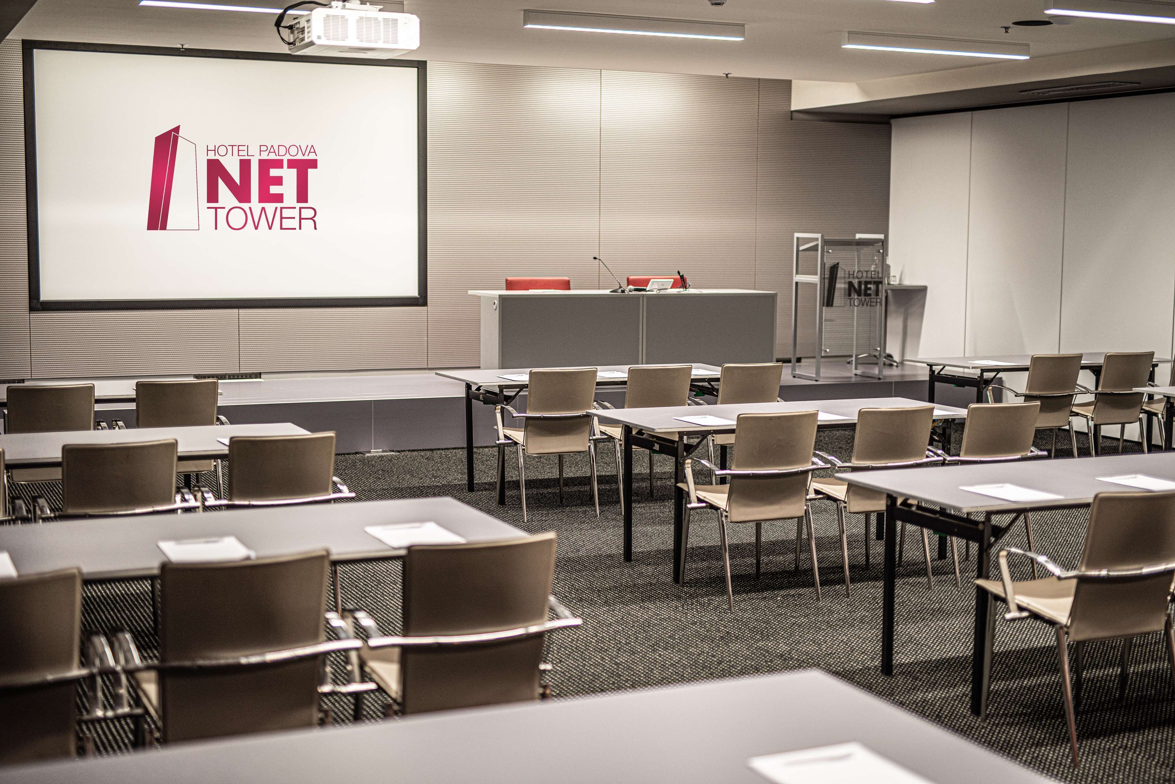 Sala de conferências com uma tela grande exibindo o logotipo Hotel Padova Net Tower, várias mesas com cadeiras e um pódio.