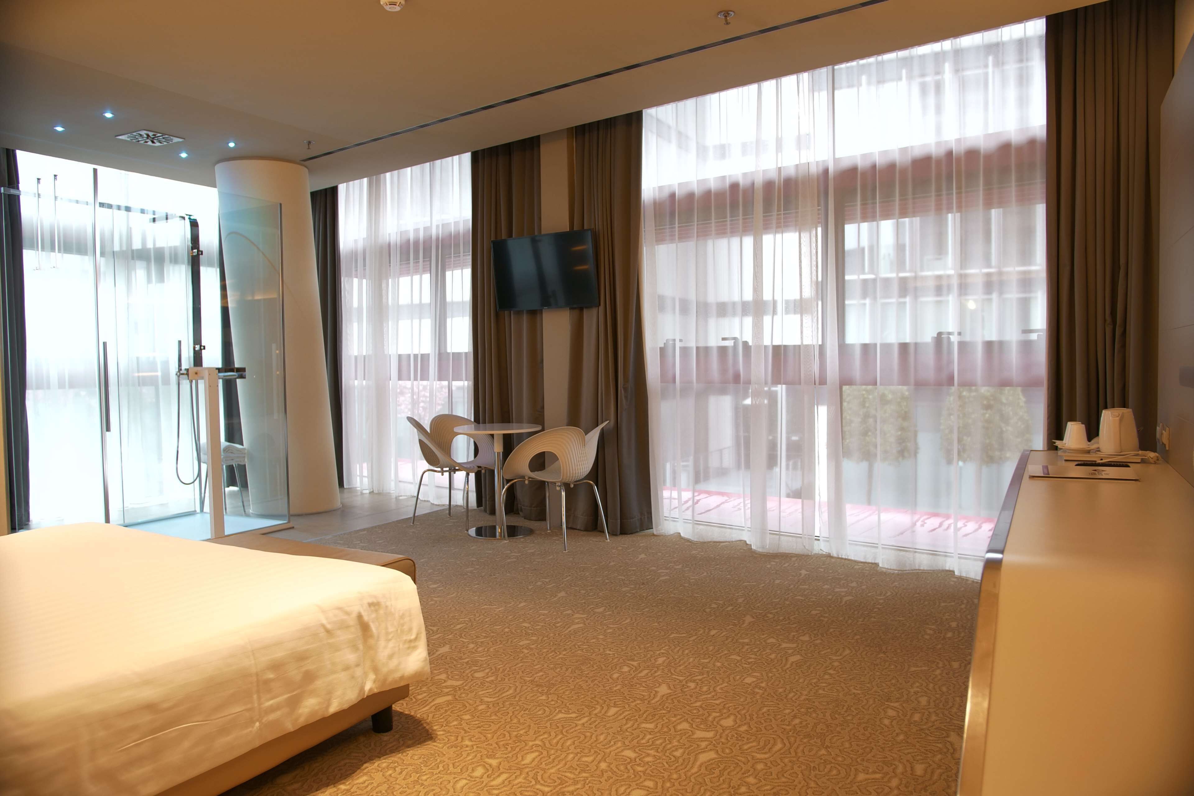 A hotel room with a bed, a desk, a TV, a shower area, and large windows with curtains.