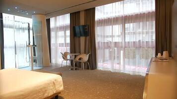 A hotel room with a bed, a desk, a TV, a shower area, and large windows with curtains.