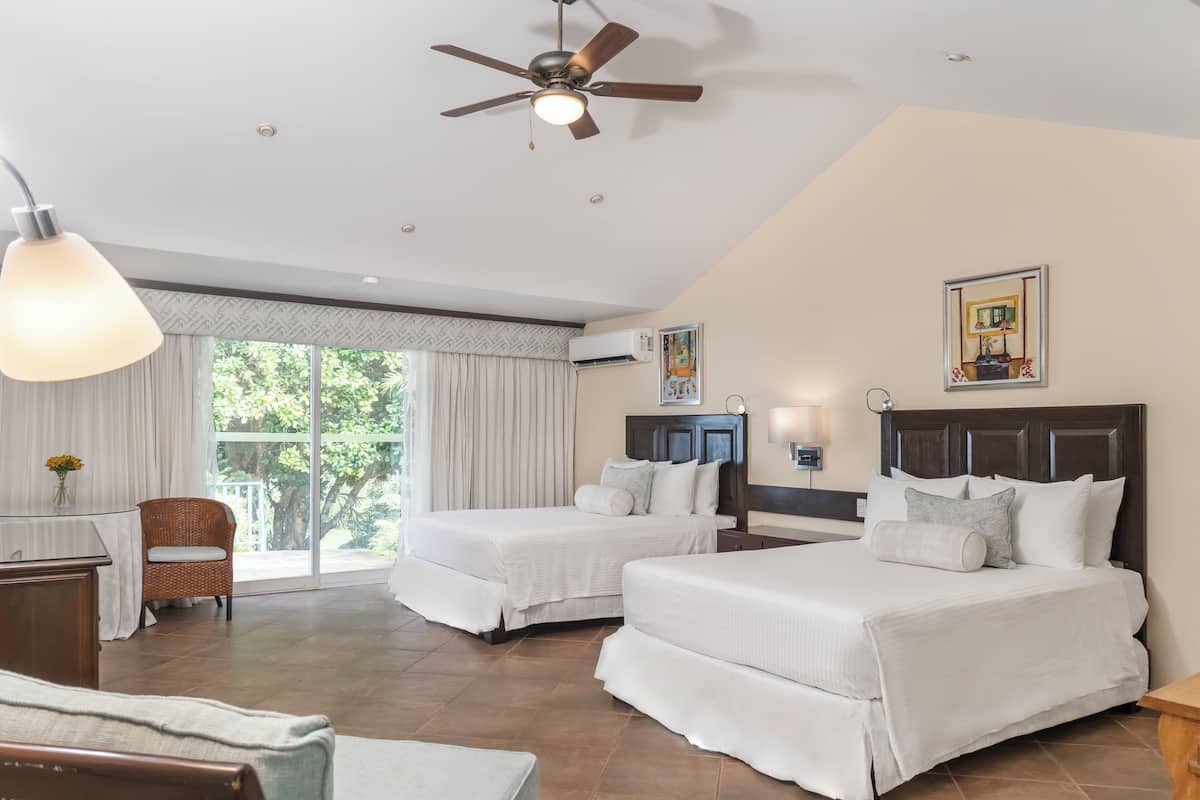 A hotel room with two beds, a desk, a chair, a ceiling fan, and a large window.
