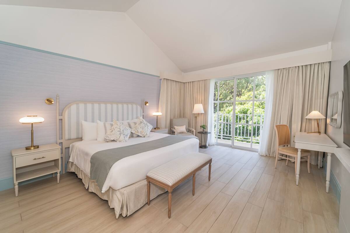 A bedroom with a bed, bedside tables, a bench, a chair, a desk, and a large window with curtains.