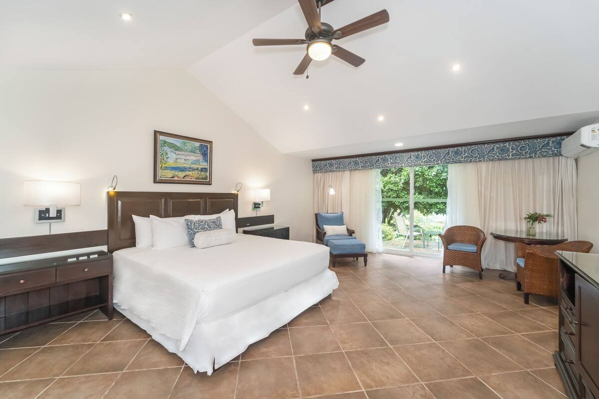 A spacious bedroom with a large bed, a ceiling fan, a desk, and a view of greenery outside.