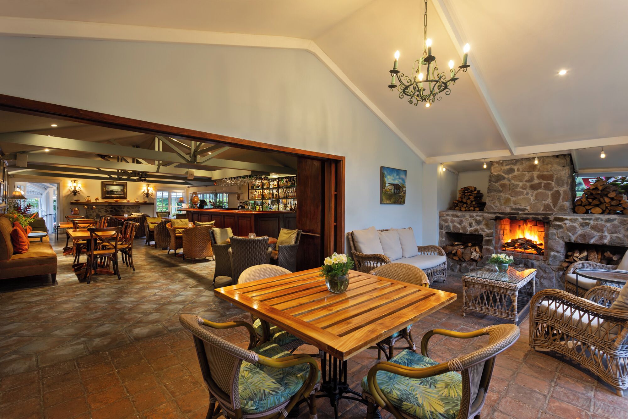 Amplia sala de estar con mesa de comedor de madera, mobiliario de mimbre, chimenea de piedra y zona de bar con vistas al exterior.