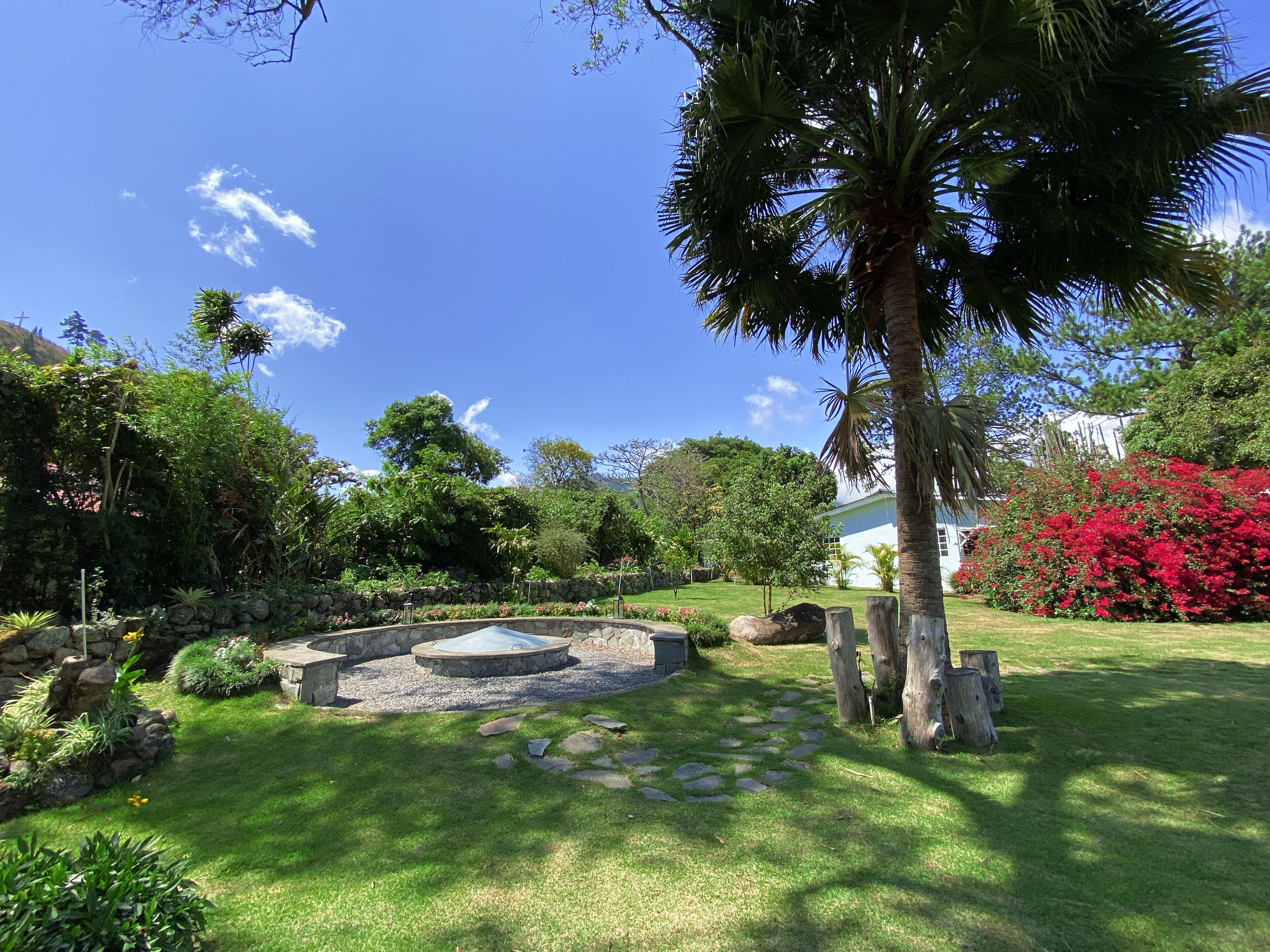 Un jardín bien cuidado con una fuente de piedra, abundante vegetación y un cielo azul despejado.