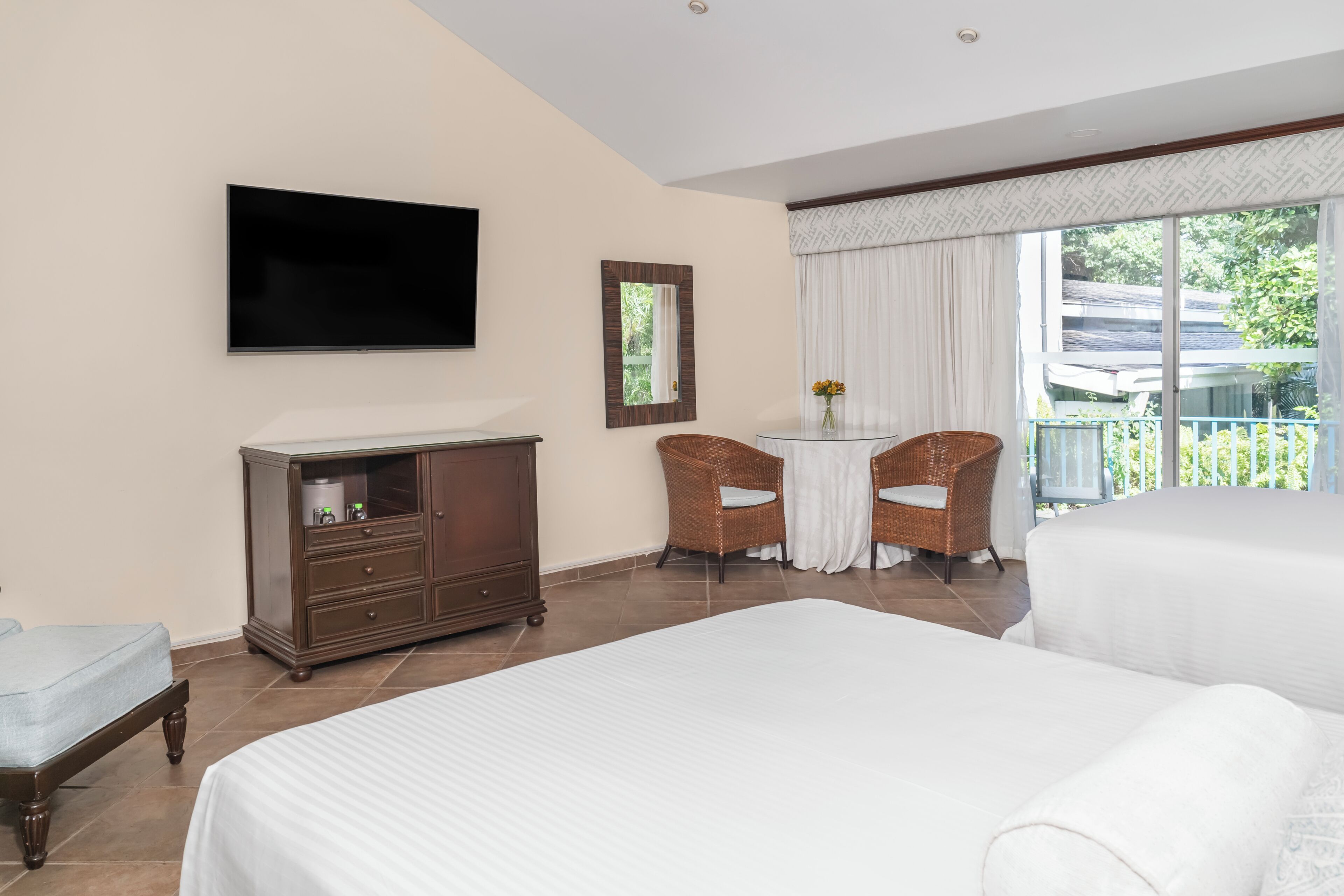 Quarto de hotel com duas camas, televisão, penteadeira, mesa pequena com cadeiras e varanda com vista.