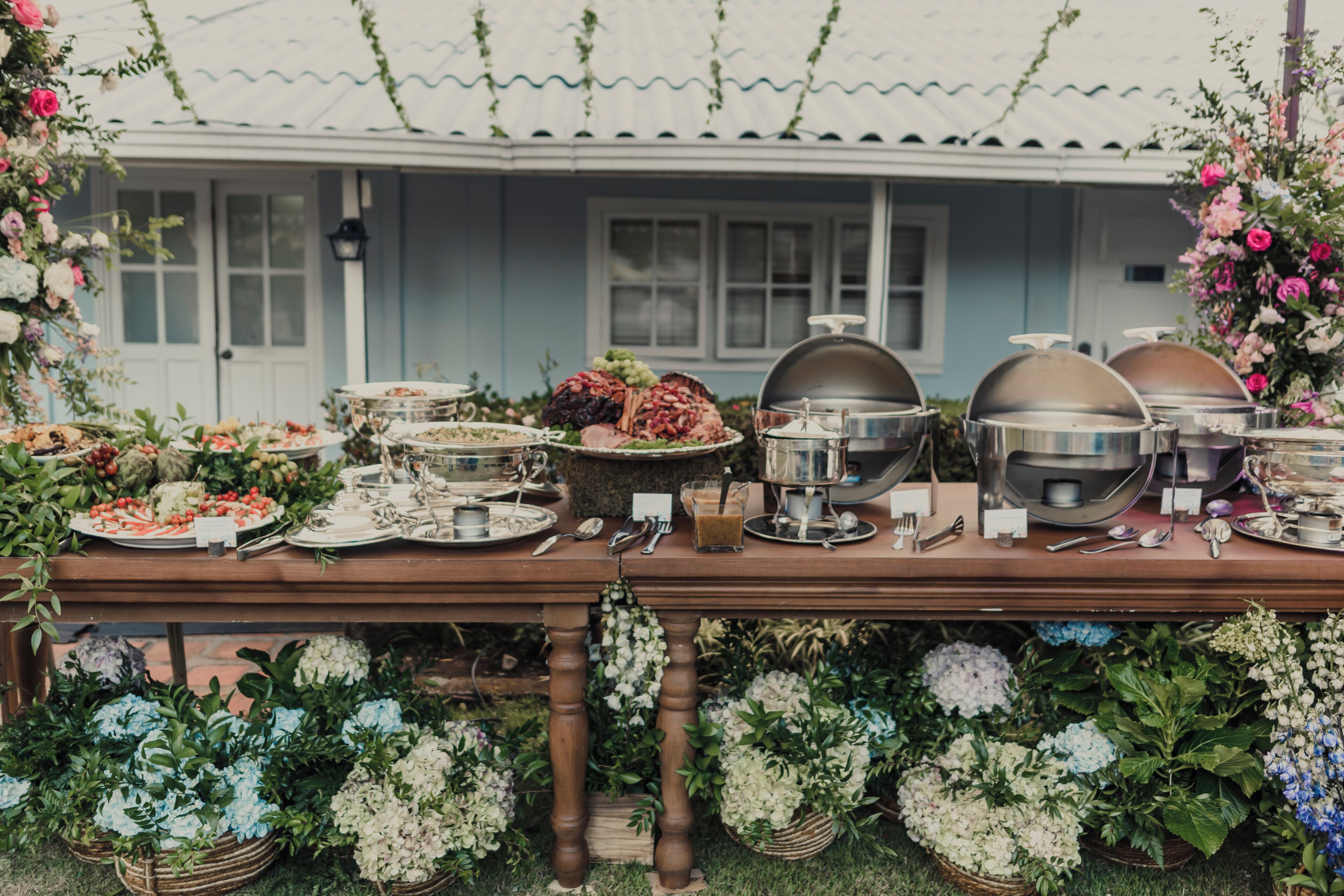 A buffet table with various dishes and drinks, set up outdoors with a house in the background.