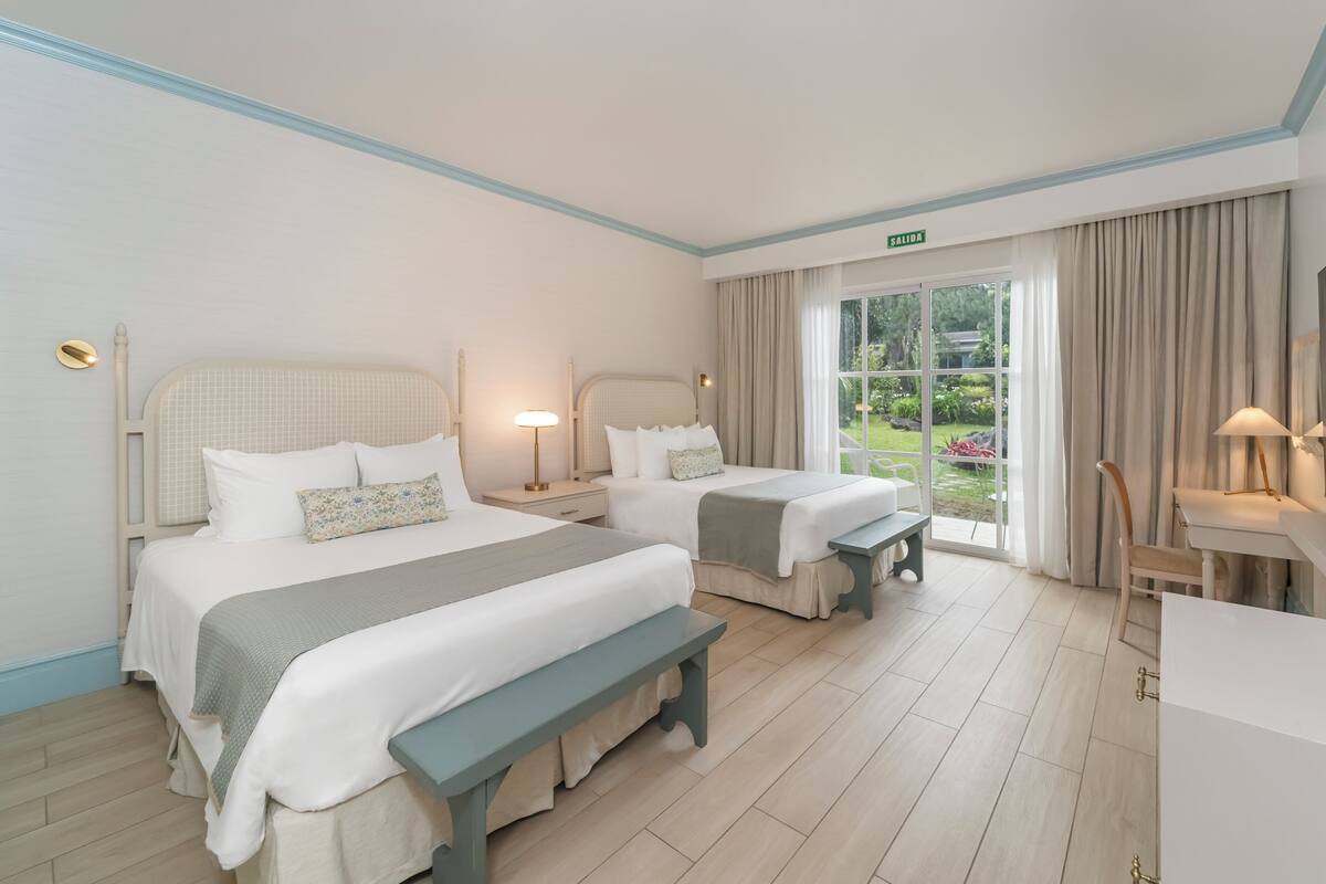 A hotel room with two beds, a bench, a desk, and a view of the garden.