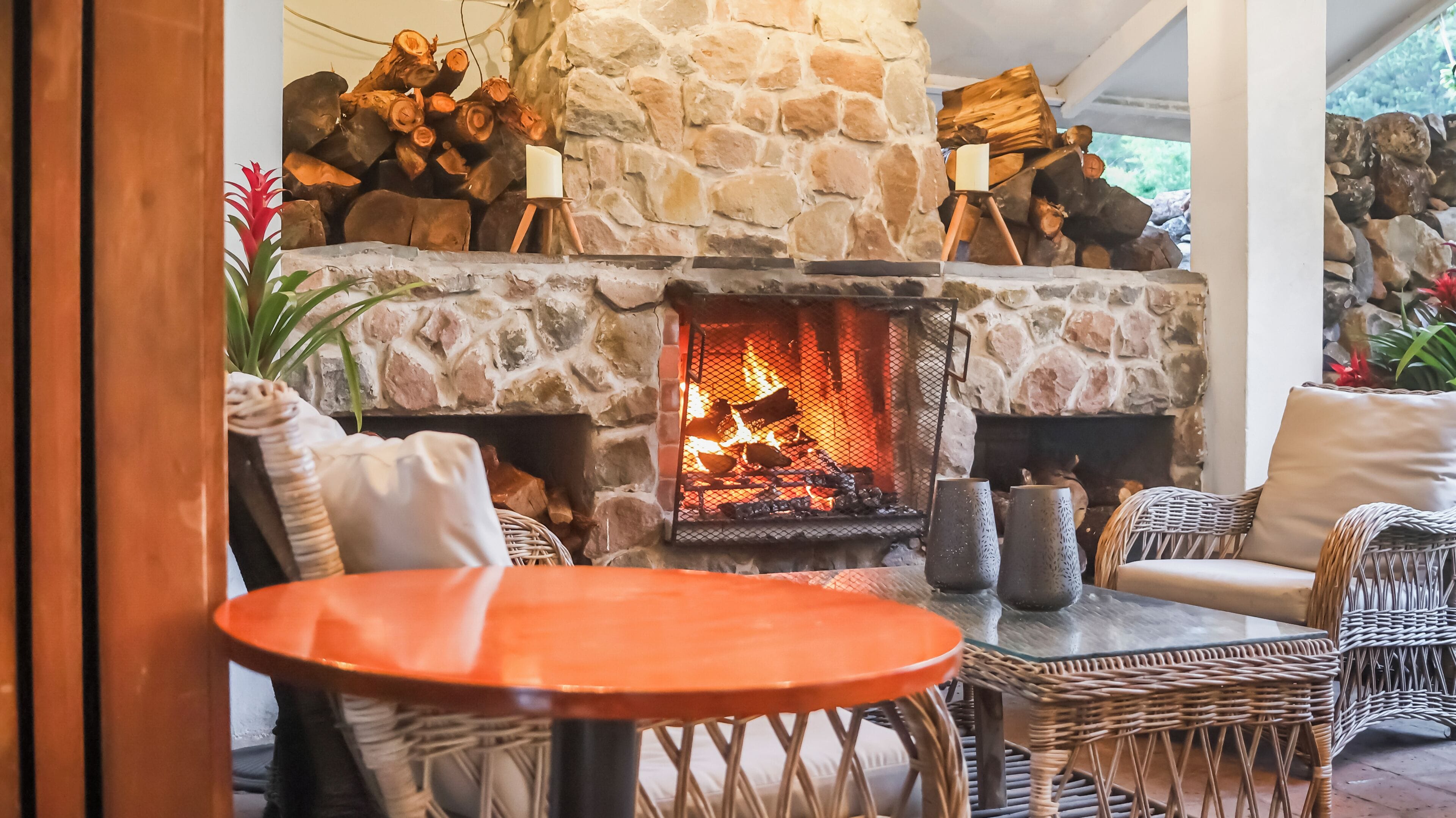 A stone fireplace with logs, wicker furniture, and a table in a cozy indoor setting.