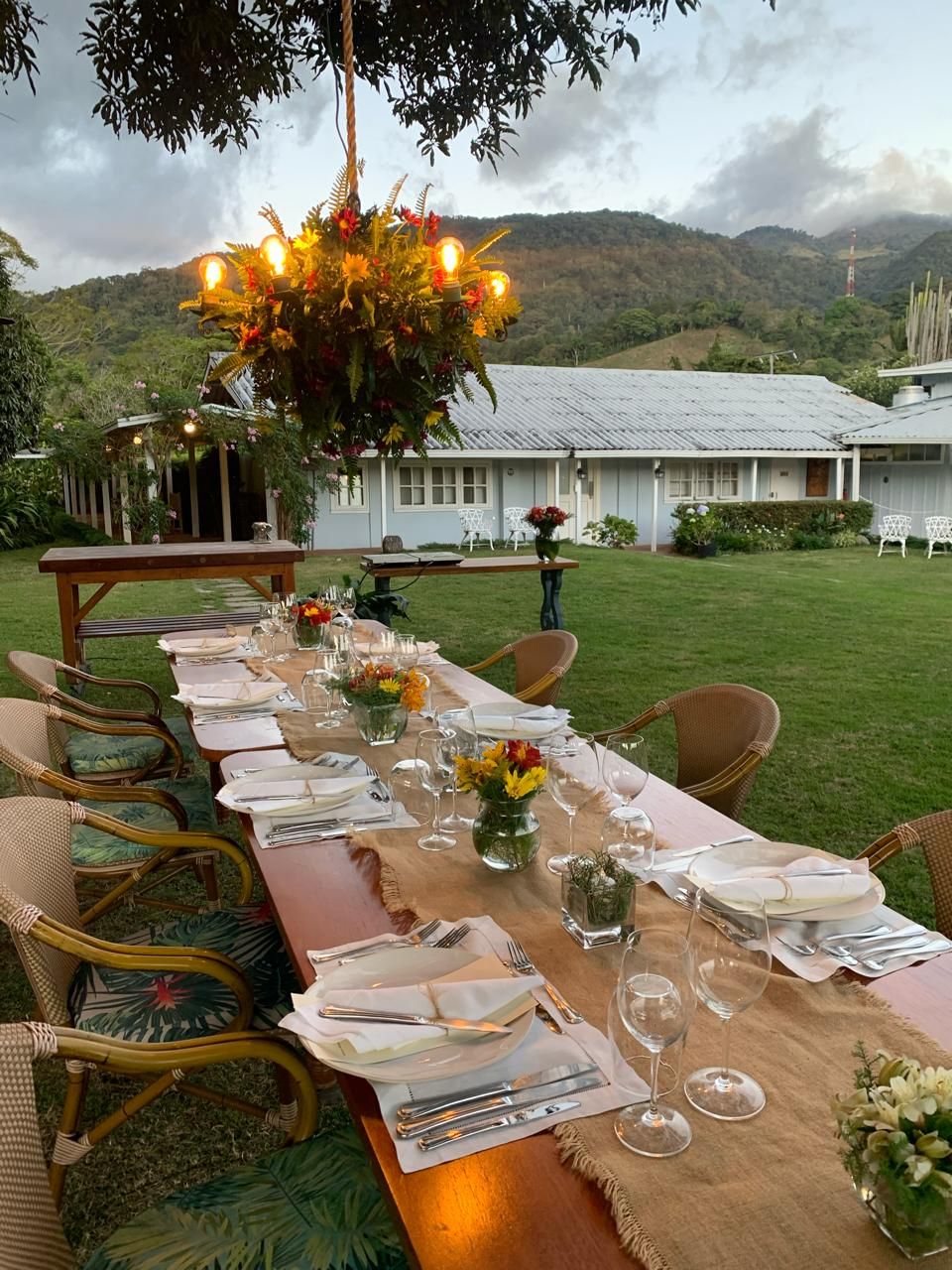 A long dining table set outdoors with chairs, a hanging light fixture, and a view of a house and hills in the background.