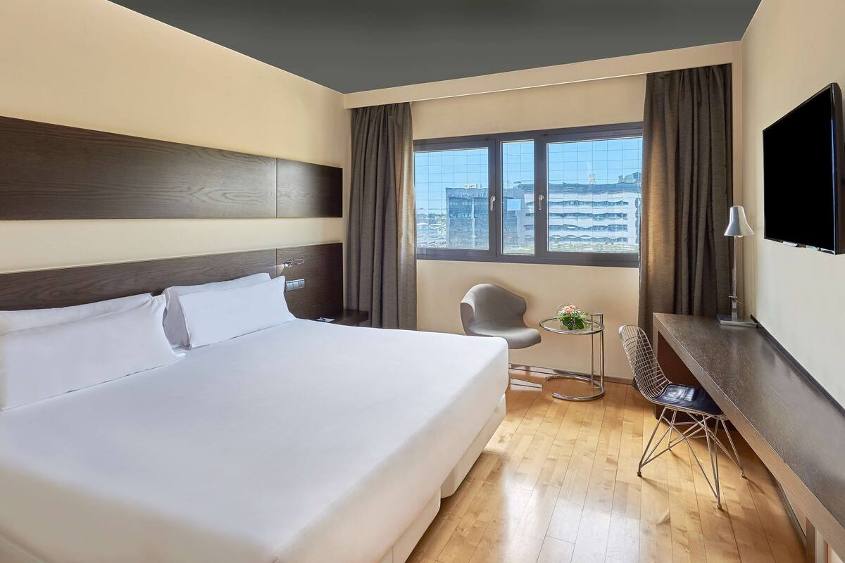 Ein Hotelzimmer mit einem großen Bett, einem Schreibtisch, einem Sessel, einem Fernseher und einem Fenster mit Blick auf die Stadt.