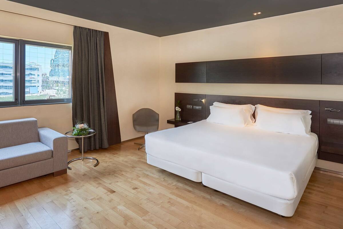 Ein modernes Hotelzimmer mit einem großen Bett, einer Couch, einem kleinen Tisch mit Blumen und Blick auf die Stadt.